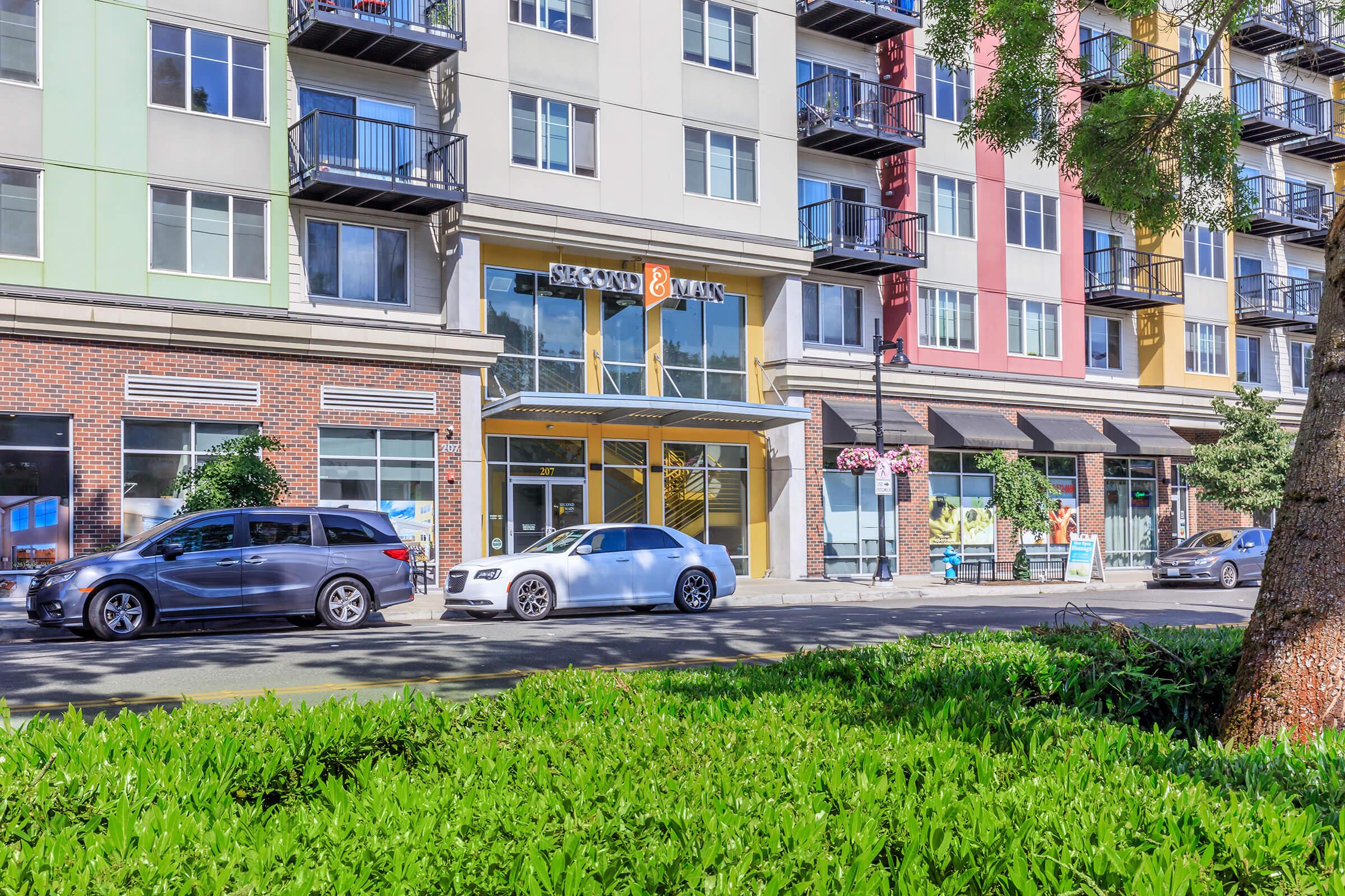 A modern multi-story apartment building with a mix of colorful facades. The ground level features large windows and a welcoming entrance. Parked cars line the street in front, and there are trees and shrubs providing greenery along the sidewalk. The scene conveys an urban residential atmosphere.