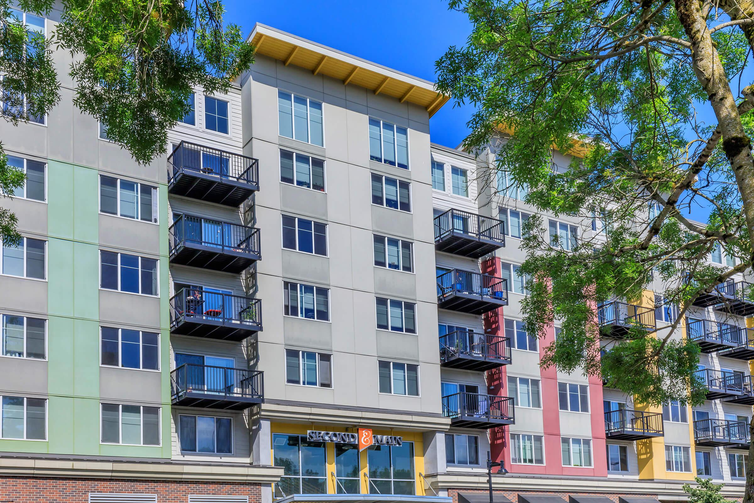 Modern apartment building with multiple stories and a mix of colorful exterior panels. Balconies line the upper floors, and the entrance features a stylish sign, surrounded by lush trees under a clear blue sky.