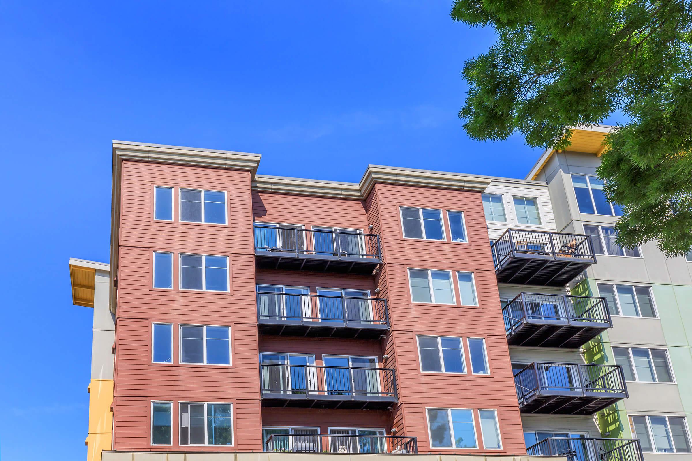 A modern apartment building with a mix of brown and gray siding. The structure features multiple balconies with black railings, large windows, and a clear blue sky in the background, along with some greenery from nearby trees.