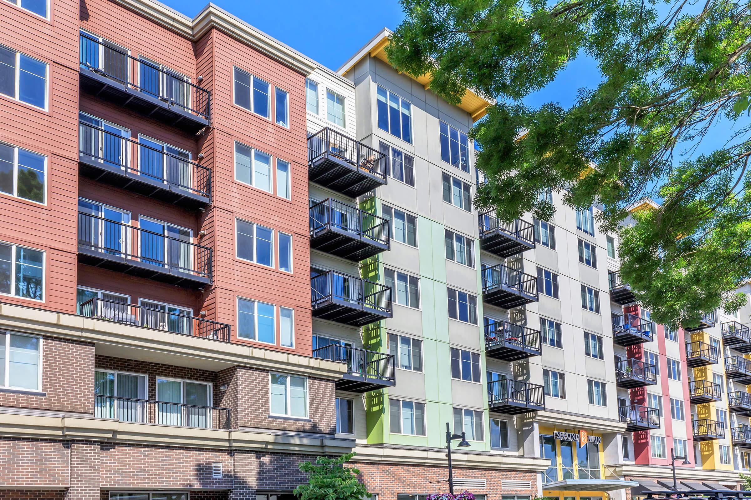 A modern apartment building with colorful facades featuring multiple balconies and large windows. The structure includes a mix of brick and siding materials, set against a clear blue sky, with trees partially framing the view.