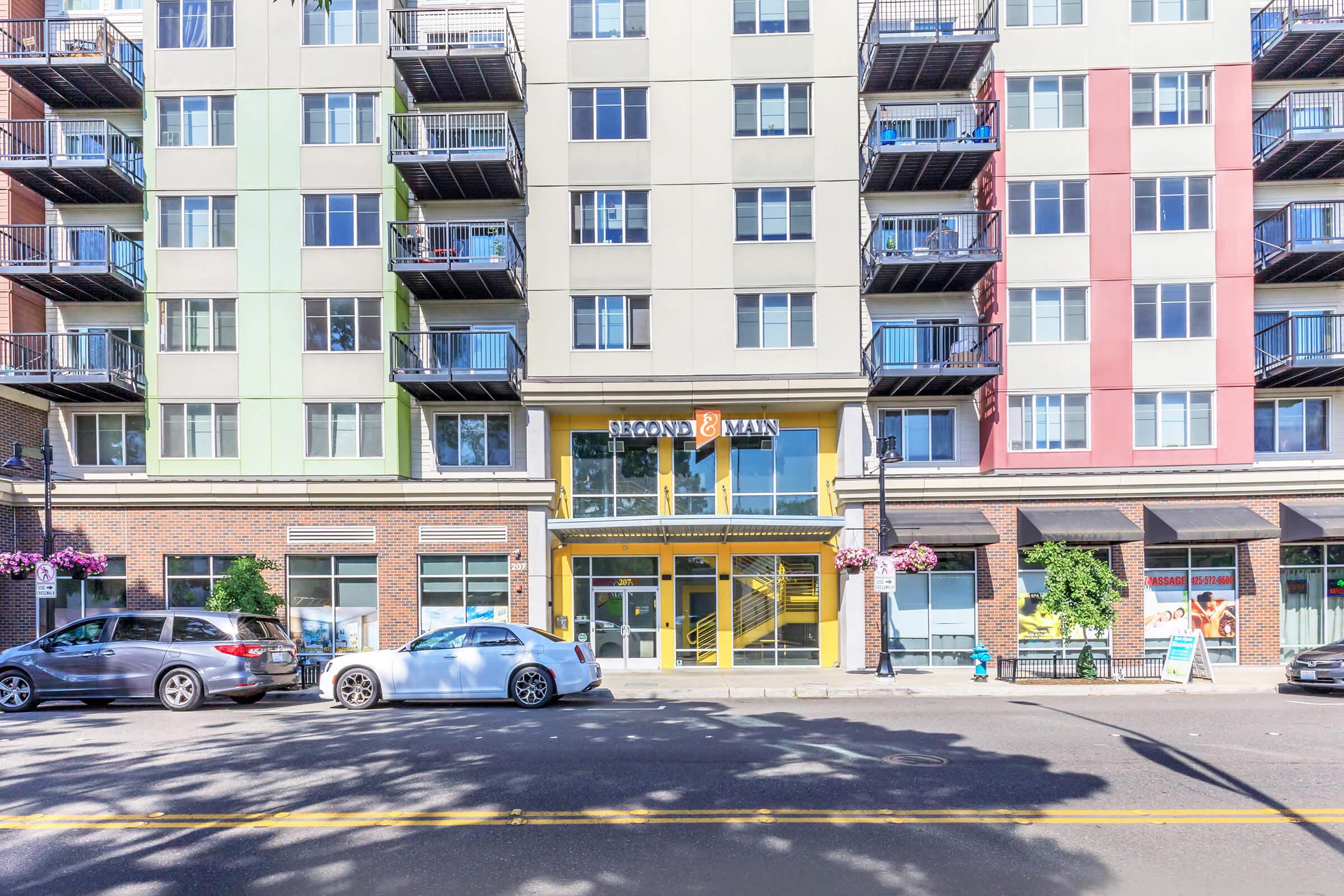 A modern apartment building with multicolored exterior panels, featuring a prominent entrance with glass doors and the name "Regan Man" displayed above. There are several balconies and flower planters along the walkway. Parked cars are seen in front of the building, along a tree-lined street.