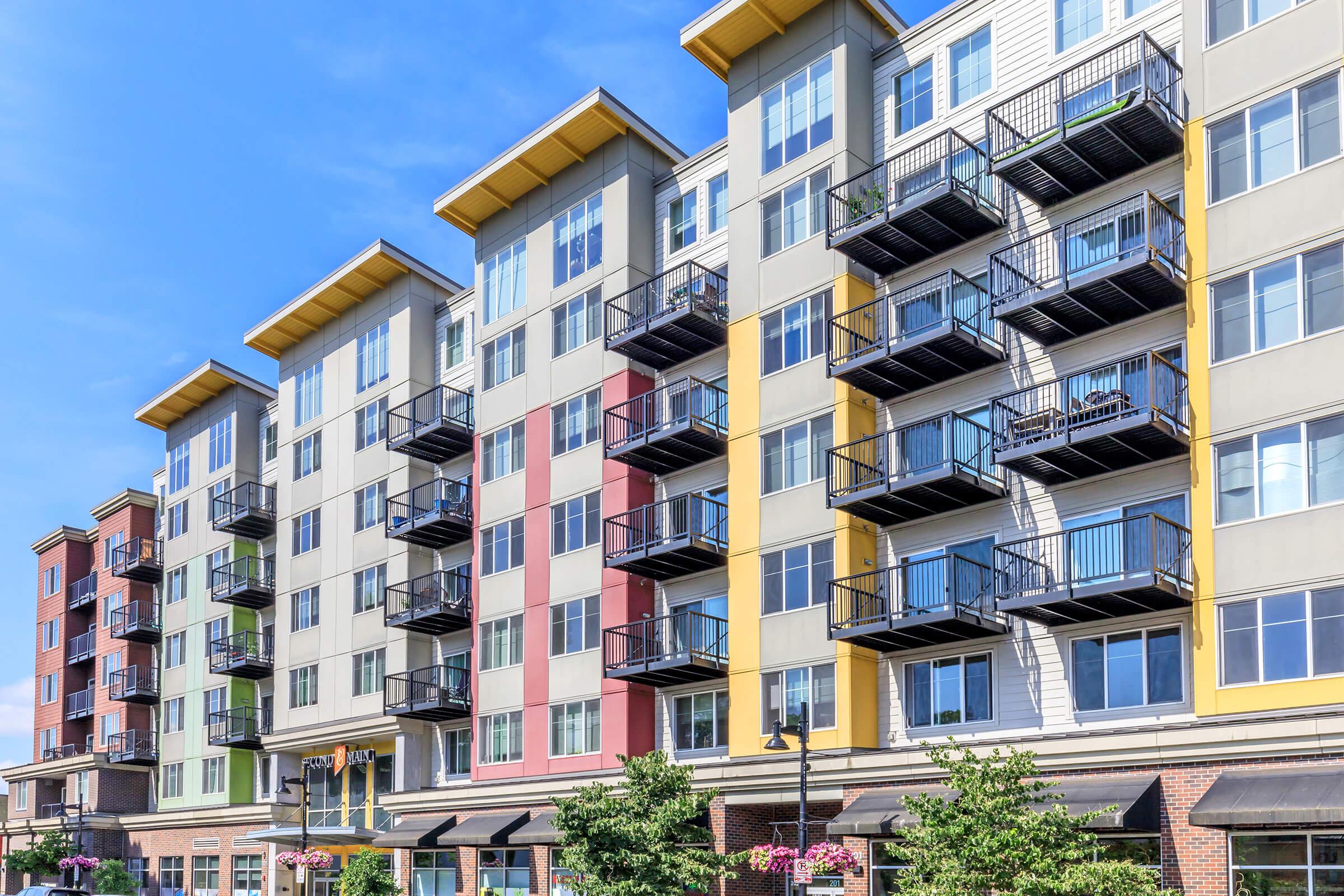 A modern apartment building with five stories featuring a mix of colorful facades in shades of yellow, green, red, and gray. Each floor has balconies and large windows, surrounded by trees and decorative plants along the ground level. The sky is clear and blue, enhancing the vibrant appearance of the structure.