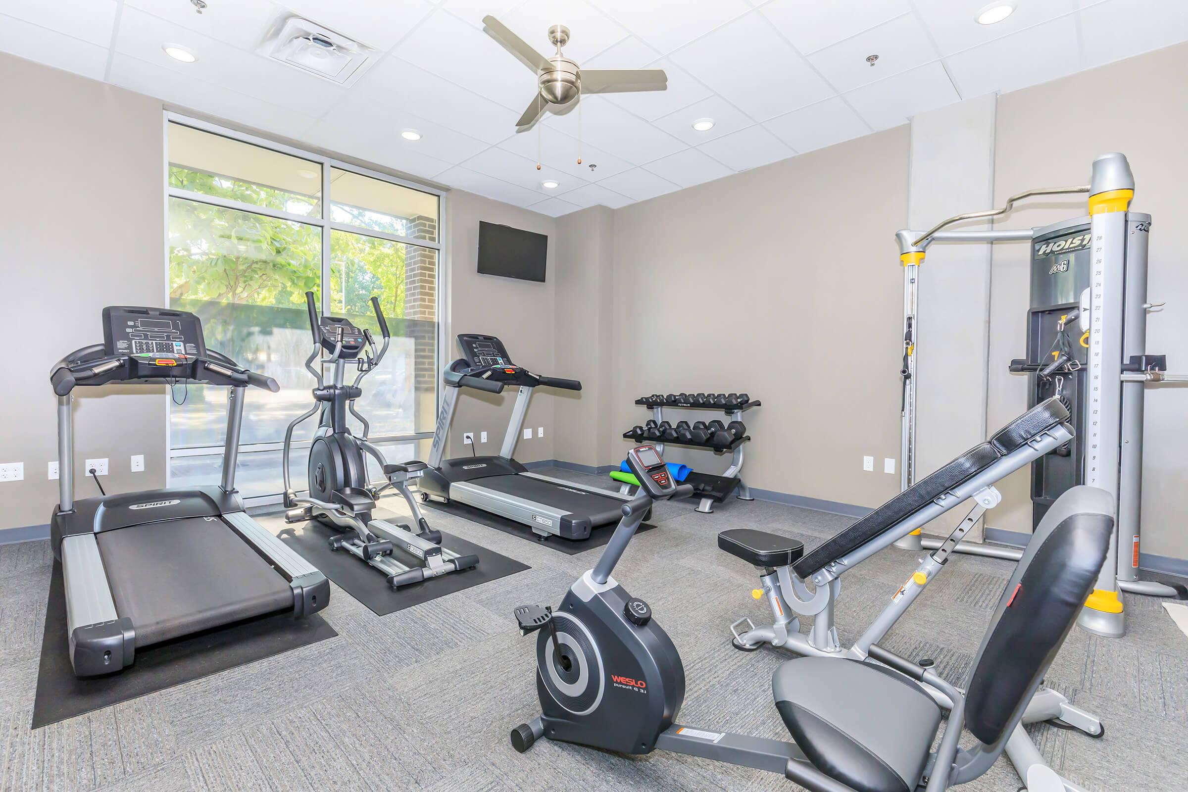 A well-equipped gym featuring two treadmills, an elliptical machine, a stationary bike, a weight bench, and a multi-station weight machine. The space has large windows allowing natural light, with gray carpet flooring and a ceiling fan for ventilation.