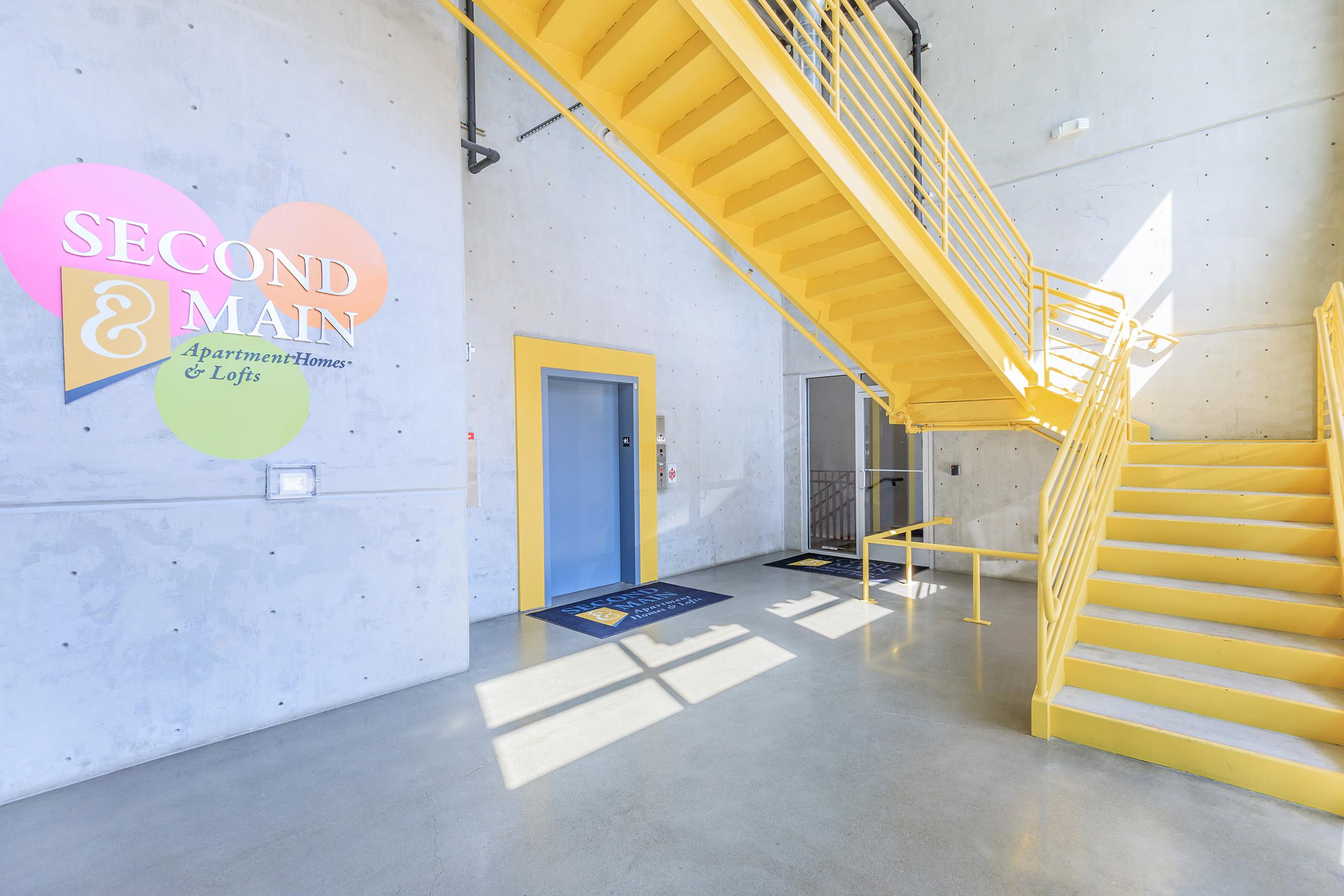 Interior view of a modern apartment building entrance with a bright yellow staircase. The wall features the text "SECOND & MAIN Apartment Homes & Lofts." There are large windows allowing natural light, and a blue mat is placed on the floor at the base of the stairs. Simple, industrial design with concrete walls.