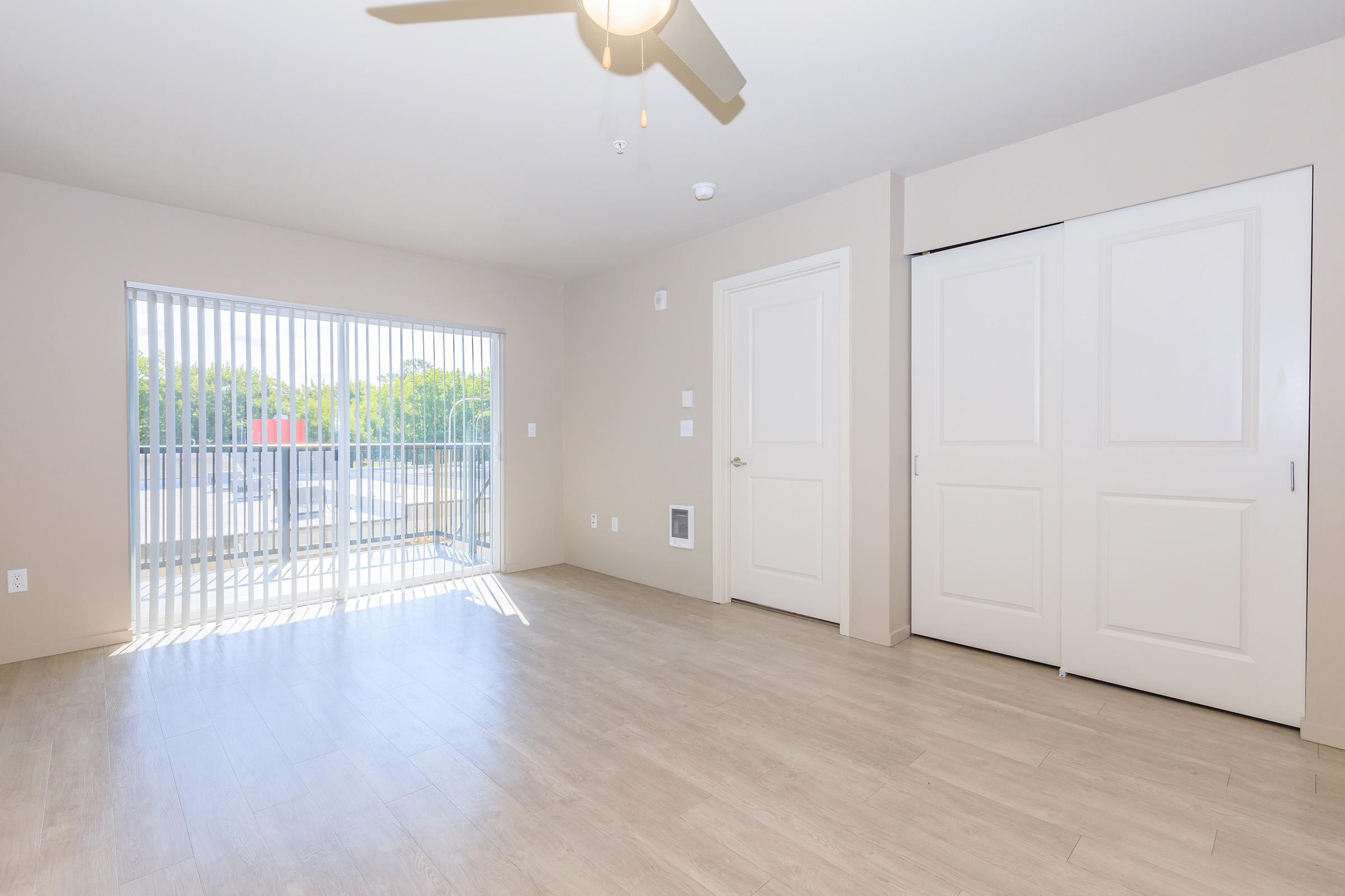 A bright and spacious empty room with light-colored walls and a wooden floor. There are large sliding doors with vertical blinds leading to a balcony, allowing natural light to fill the space. A ceiling fan is mounted above, and a closet door is visible on the right.