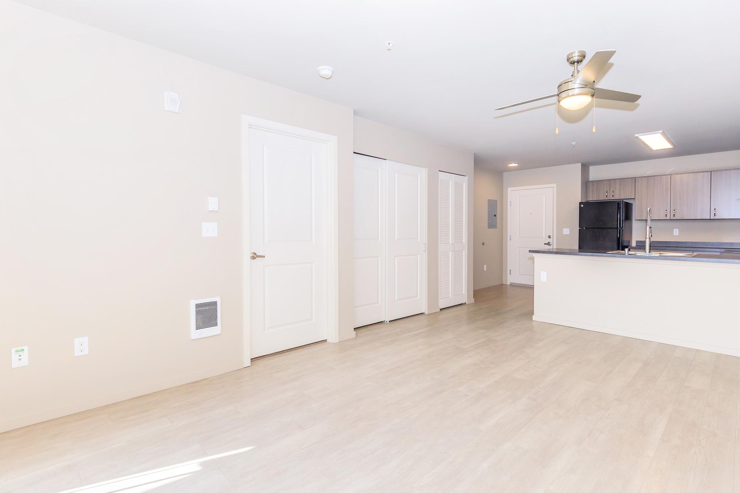 A spacious, modern living area featuring light-colored walls, a ceiling fan, and a kitchen area in the background. The room has a neutral color palette with light flooring and includes a door and closet on the left. Brightly lit with natural light, creating an inviting atmosphere.