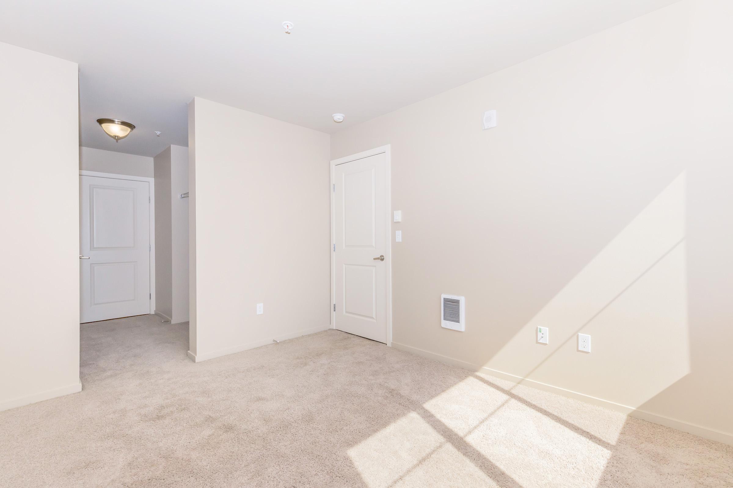 A spacious, empty room with light beige walls and carpet. The room features a closed door leading to a hallway, a ceiling light fixture, and a wall vent. Natural light streams in from a window, creating a bright and inviting atmosphere. No furniture or decorations are present.