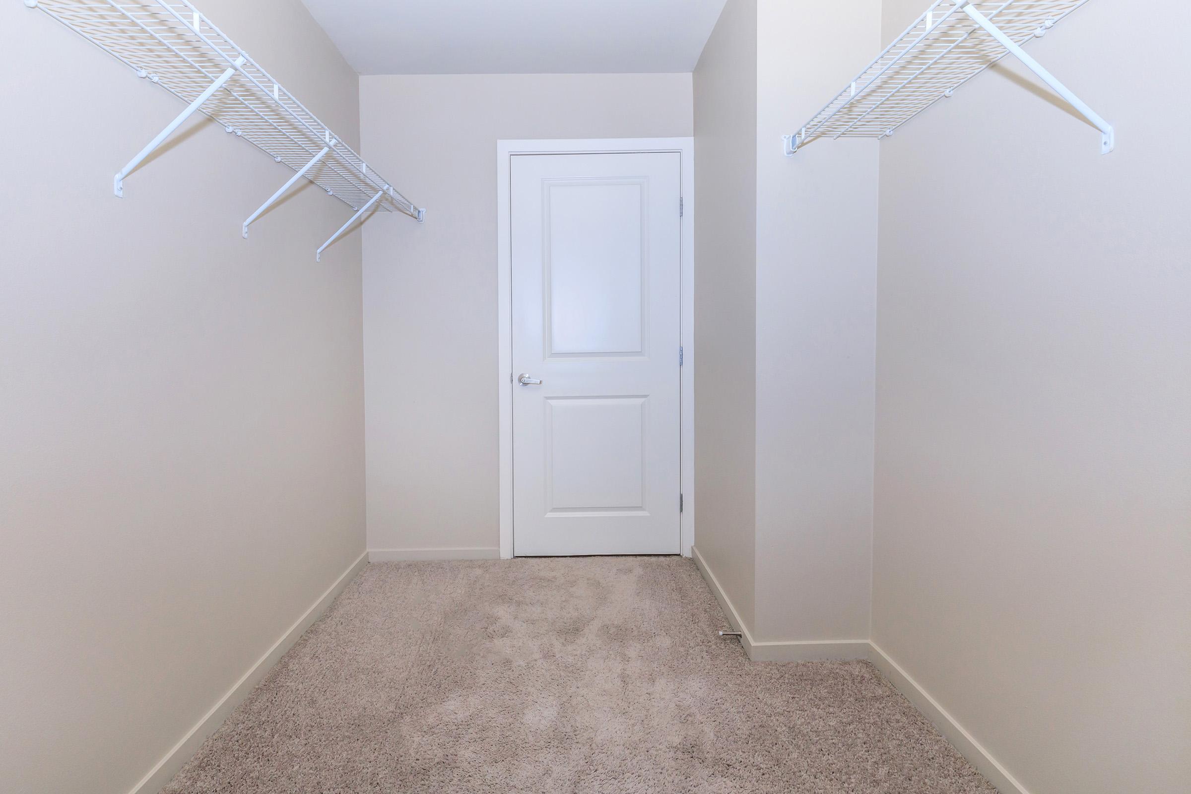 A vacant walk-in closet with light beige walls and carpet. Two wire shelving units are mounted on opposite walls, and a closed white door is visible at the far end of the room. The space appears clean and organized, ready for storage or personal items.