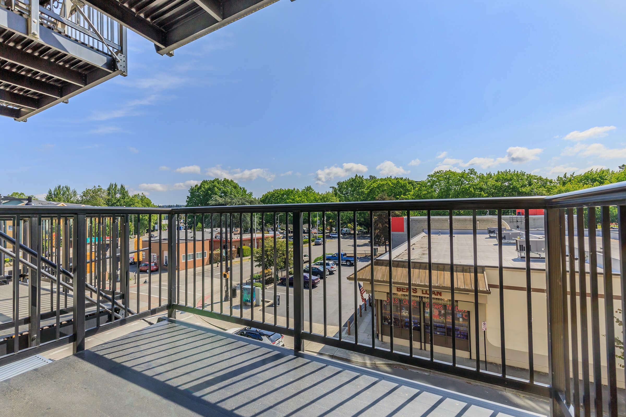 View from a balcony overlooking a parking lot and commercial area. The scene features trees in the background, a clear blue sky, and a few clouds. There are several parked cars and retail buildings visible beneath the balcony.