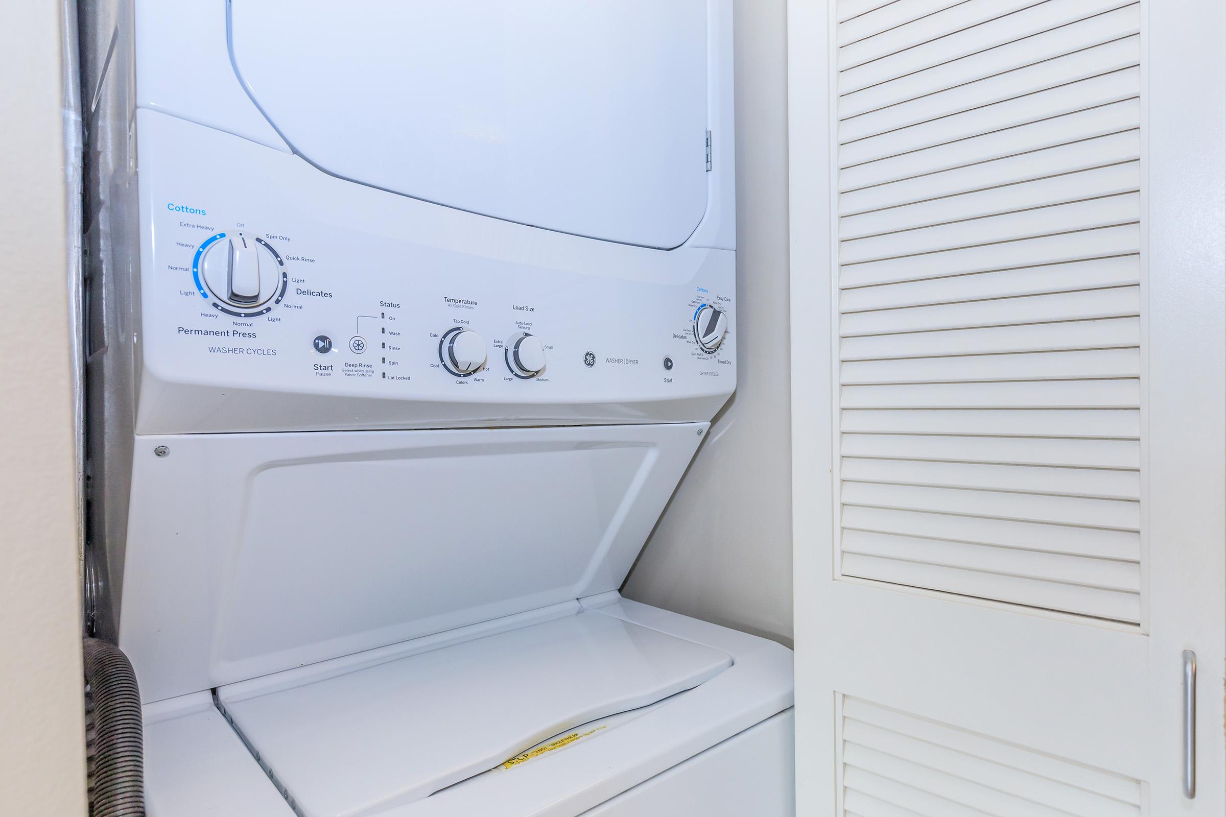 A stacked washer and dryer unit in a small laundry space, featuring a white exterior and control knobs for different wash settings, placed next to a white louvered door.