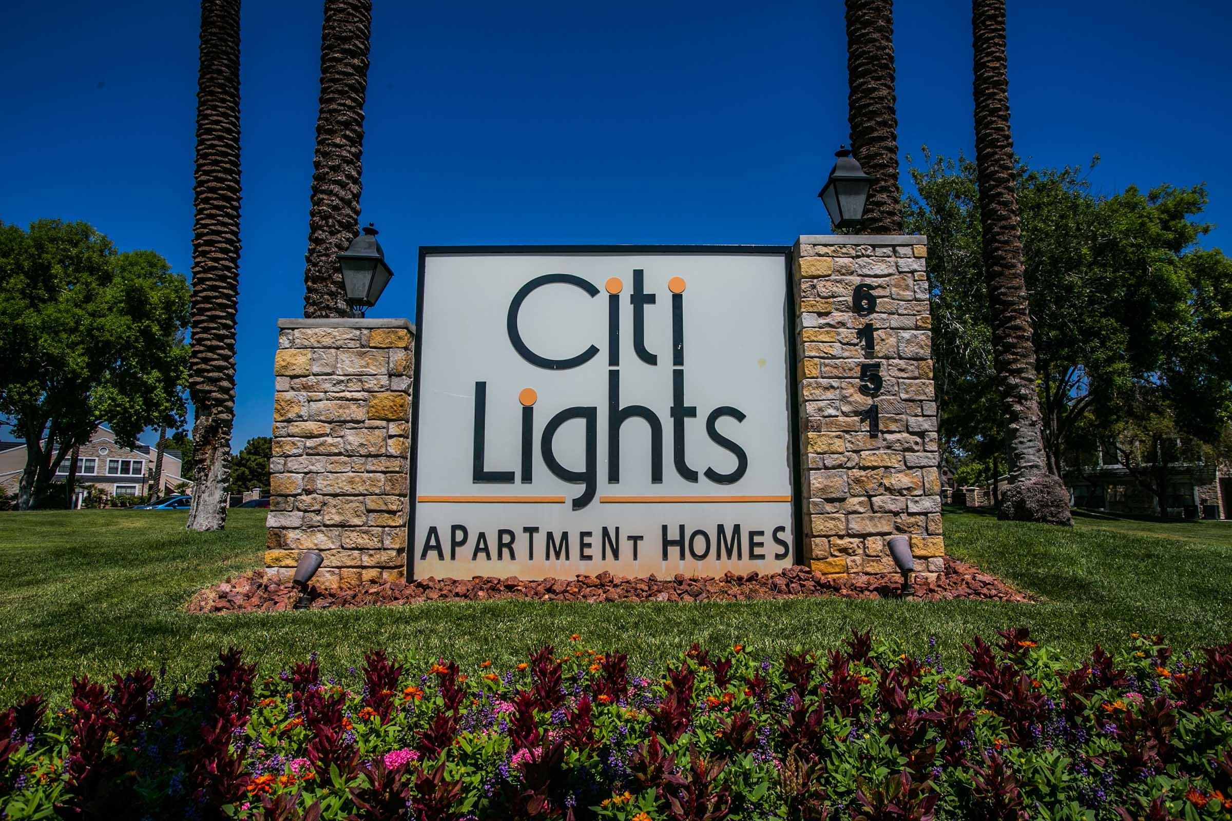 Sign for Citi Lights Apartment Homes, featuring the logo prominently displayed. The sign is set against a backdrop of green grass and colorful flowers, with palm trees and clear blue skies visible in the background. The address 6151 is also noted on the sign.