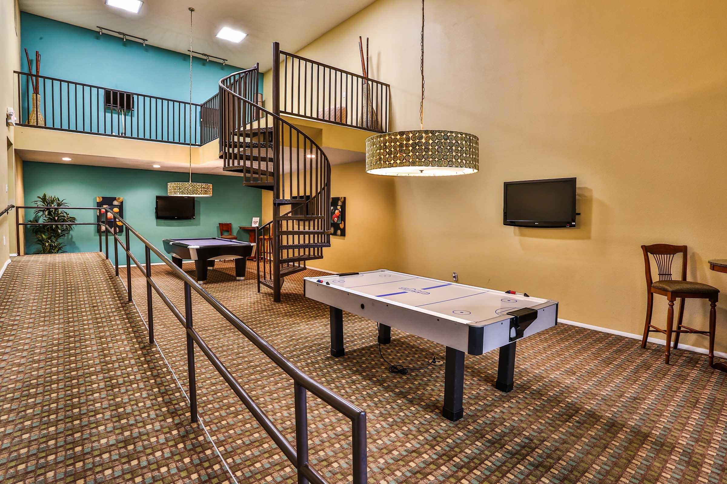 A spacious game room featuring a white air hockey table, a staircase leading to a second level, a large decorative light fixture, and a TV mounted on the wall. The walls are painted in a mix of colors, with a patterned carpet covering the floor, and there are seating options along the railing.