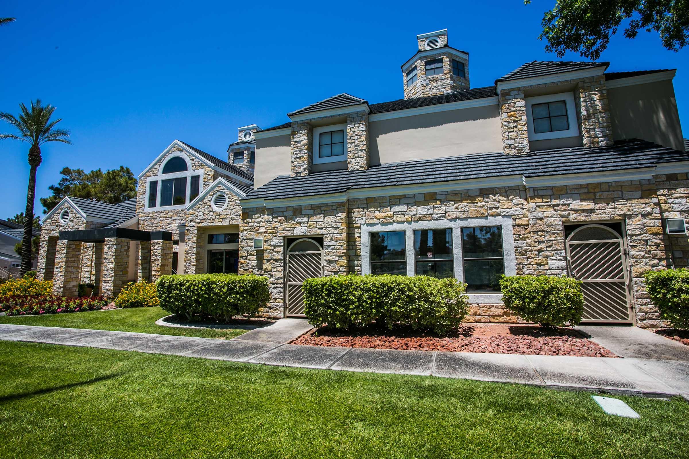 A charming two-story stone building with a steep roof, featuring large windows and a welcoming entrance. Lush green grass and neatly trimmed bushes surround the property, along with colorful flower beds. Palm trees are visible nearby under a clear blue sky.