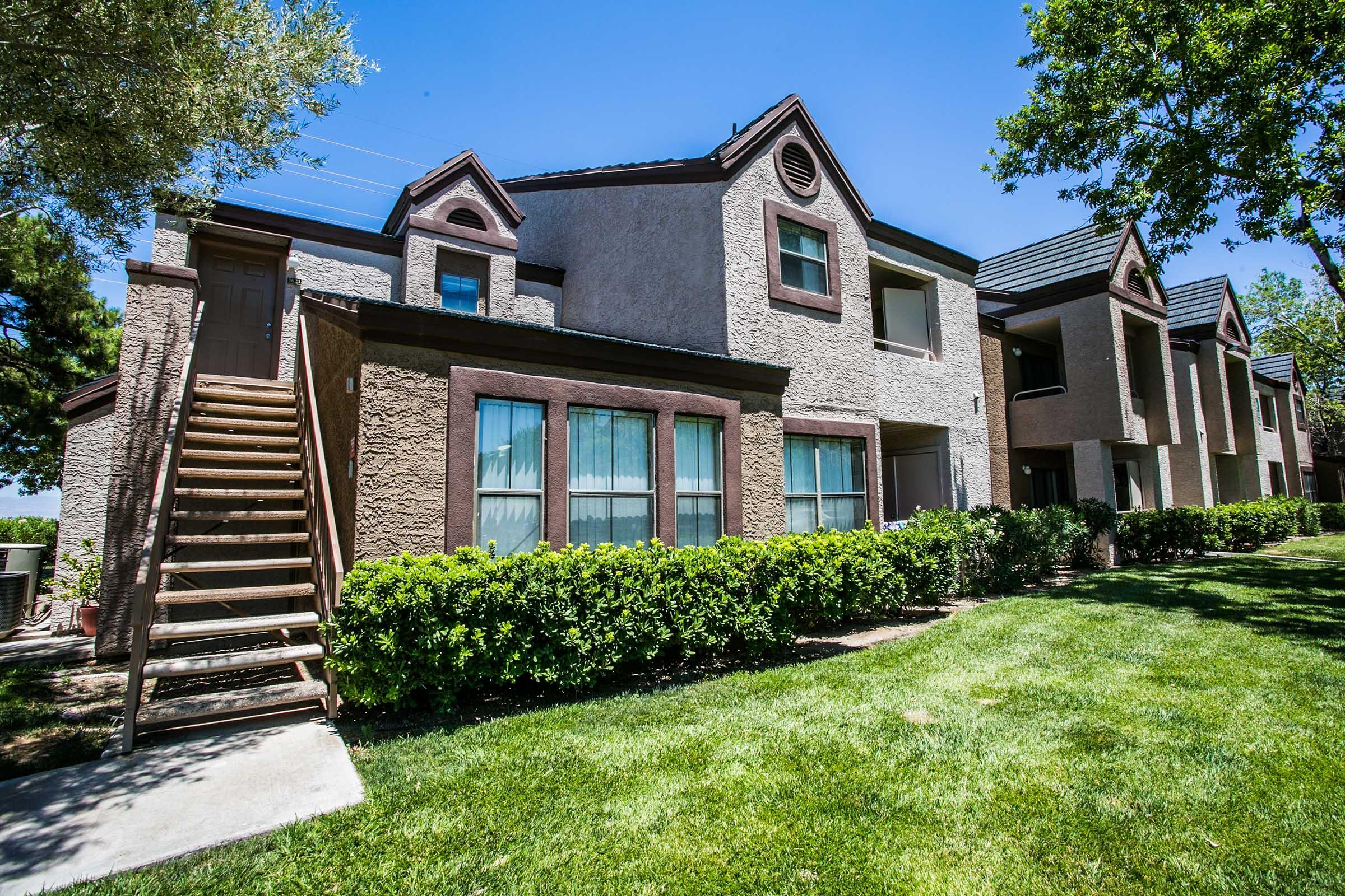 A two-story apartment building with beige stucco and brown accents, featuring multiple windows and a wooden staircase on the left. The surrounding area includes well-maintained green lawns and bushes under a clear blue sky.