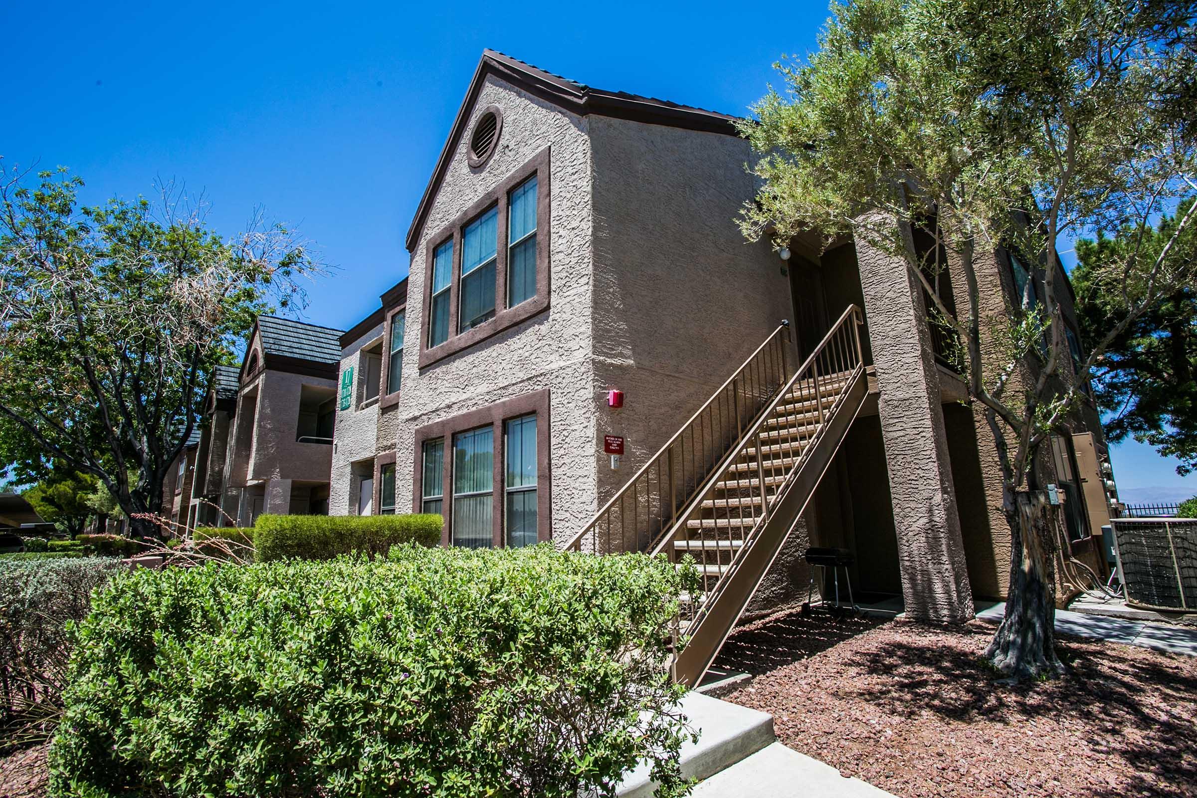 A two-story apartment building with beige exterior, featuring multiple windows and a staircase leading up to the entrance. The surrounding area includes landscaped bushes and trees, under a clear blue sky. The scene conveys a welcoming residential atmosphere.