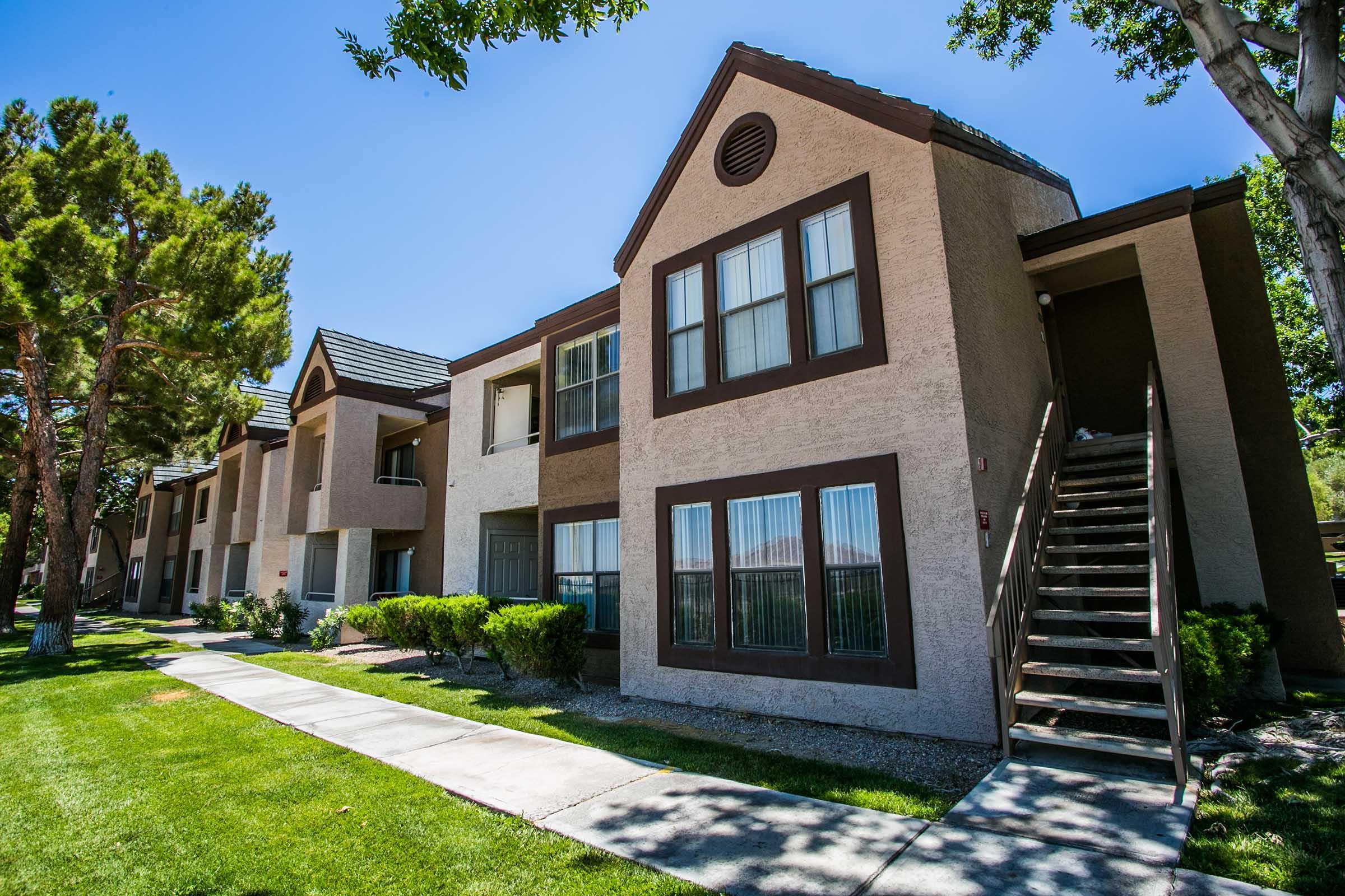 A modern apartment complex featuring two-story buildings with light-colored exterior and dark trim. The building has large windows, a flight of stairs leading to the second floor, and is surrounded by well-maintained grass and trees under bright blue skies.