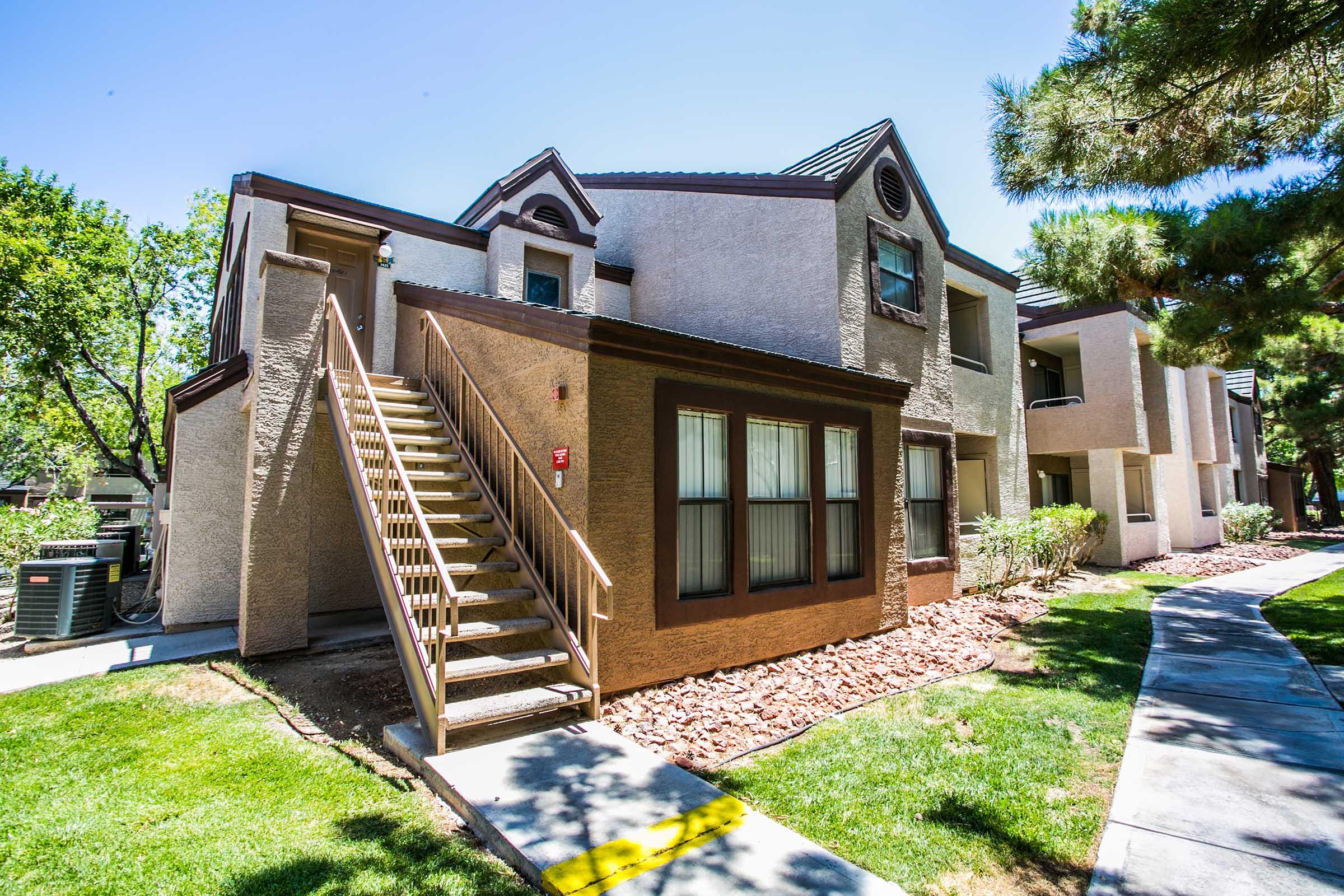 Exterior view of a two-story apartment building with a light tan facade, featuring a wooden staircase leading to the second floor. The building is surrounded by well-maintained lawns and shrubs, under a clear blue sky.