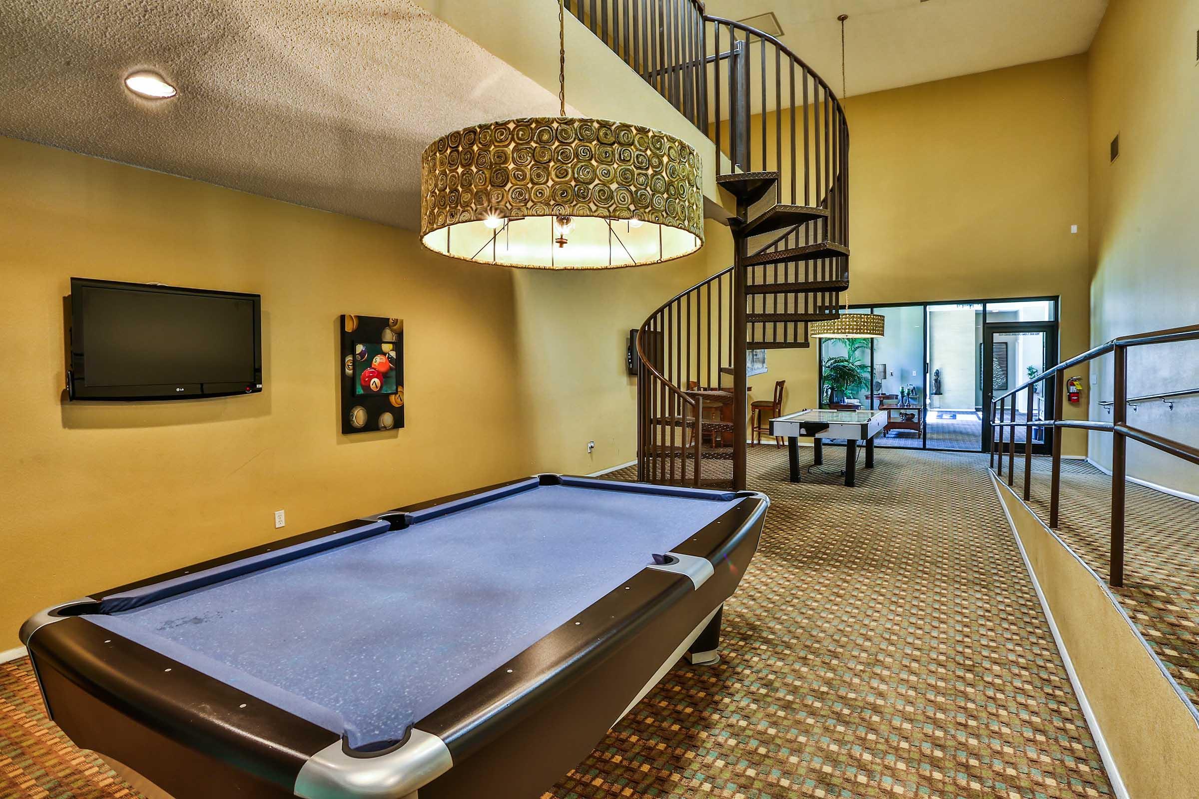 A spacious room featuring a pool table, a large chandelier, and a spiral staircase. The walls are painted a warm yellow, and there is a flat-screen TV mounted on one wall. Natural light enters through large windows, and a separate dining area with a table is visible in the background.