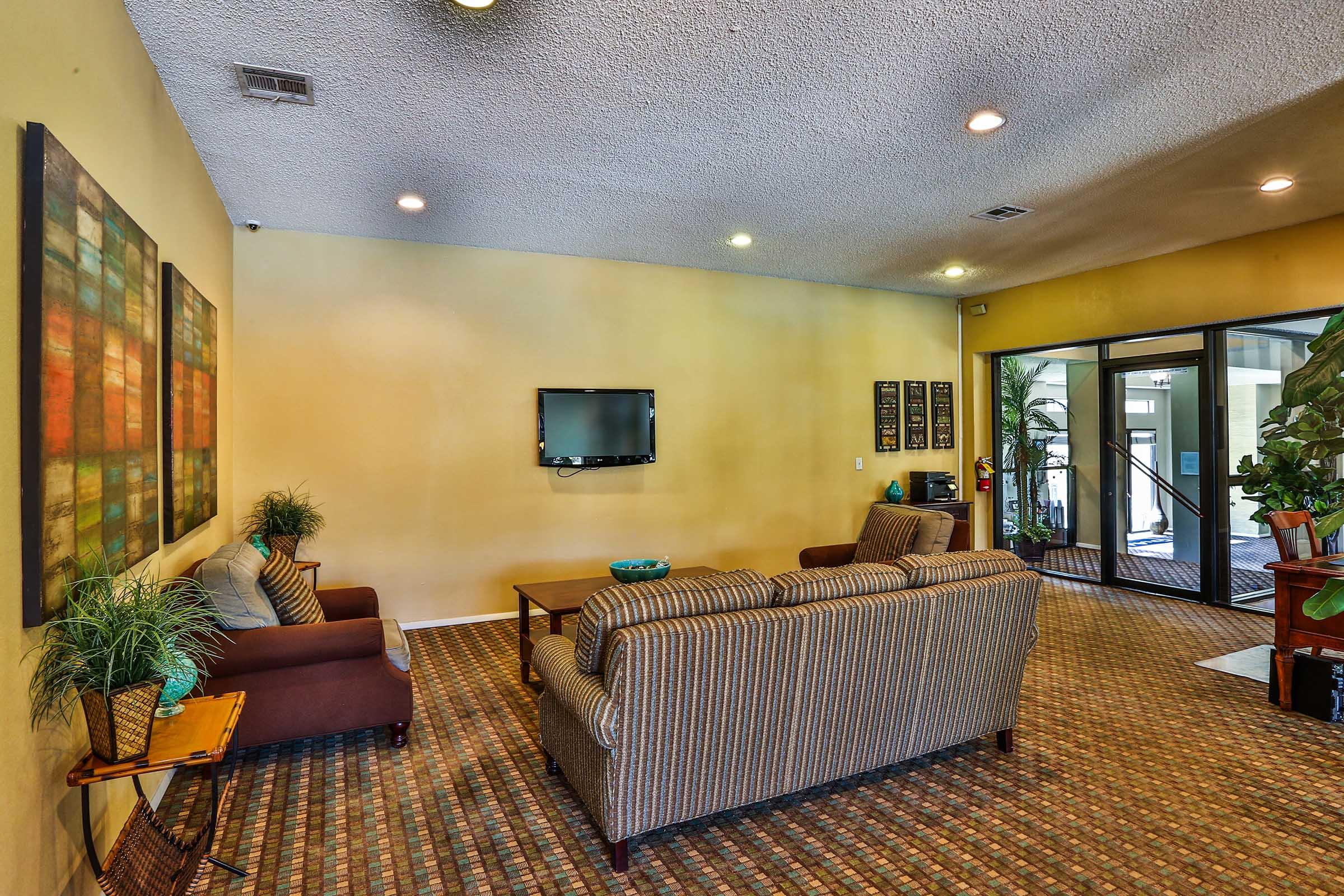 Cozy lounge area featuring two sofas, a television on the wall, and decorative artwork. There are potted plants and a welcoming atmosphere, with large windows providing natural light. The carpet has a patterned design, contributing to the relaxed ambiance of the space.