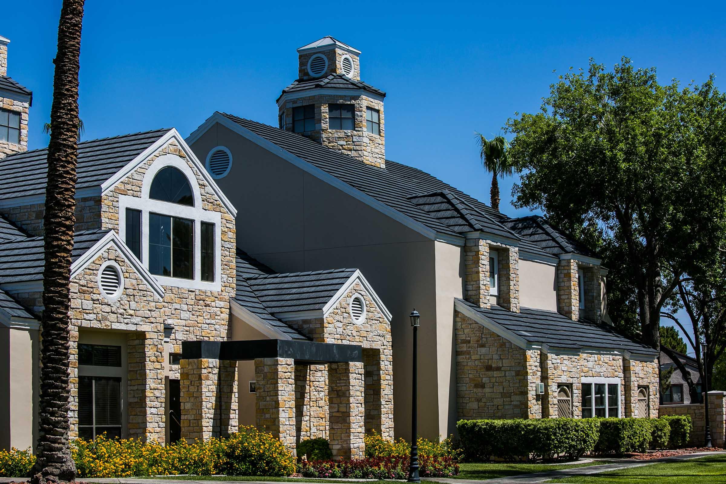A modern stone building with a unique architectural design, featuring a tower, multiple gable roofs, large windows, and a landscaped foreground with colorful flowers and palm trees under a clear blue sky.