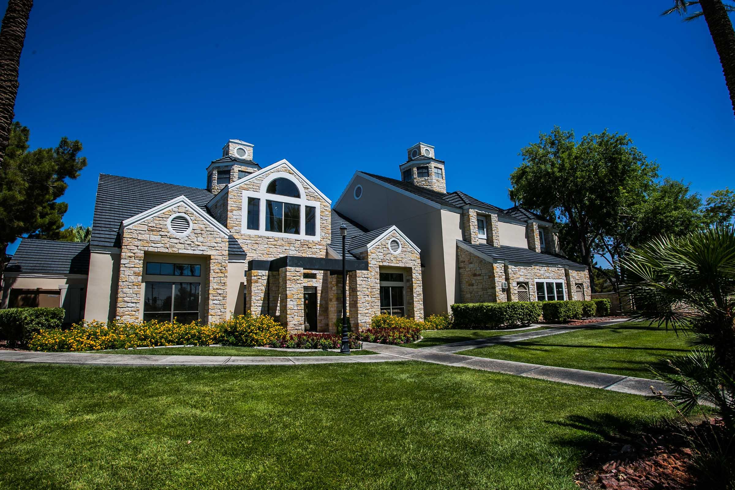 A well-maintained stone and stucco house with a large front yard, featuring neatly trimmed grass and colorful flower beds. The building has multiple gables and decorative chimneys under a clear blue sky, surrounded by lush greenery and palm trees.