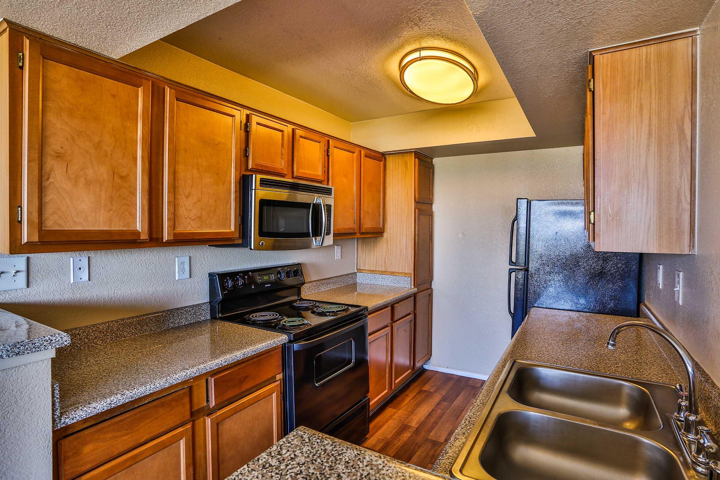 Modern kitchen with wooden cabinetry, granite countertops, and stainless steel appliances. Features include a double sink, a black oven and stove, a microwave, and a refrigerator. The flooring is hardwood, and there is overhead lighting illuminating the space.