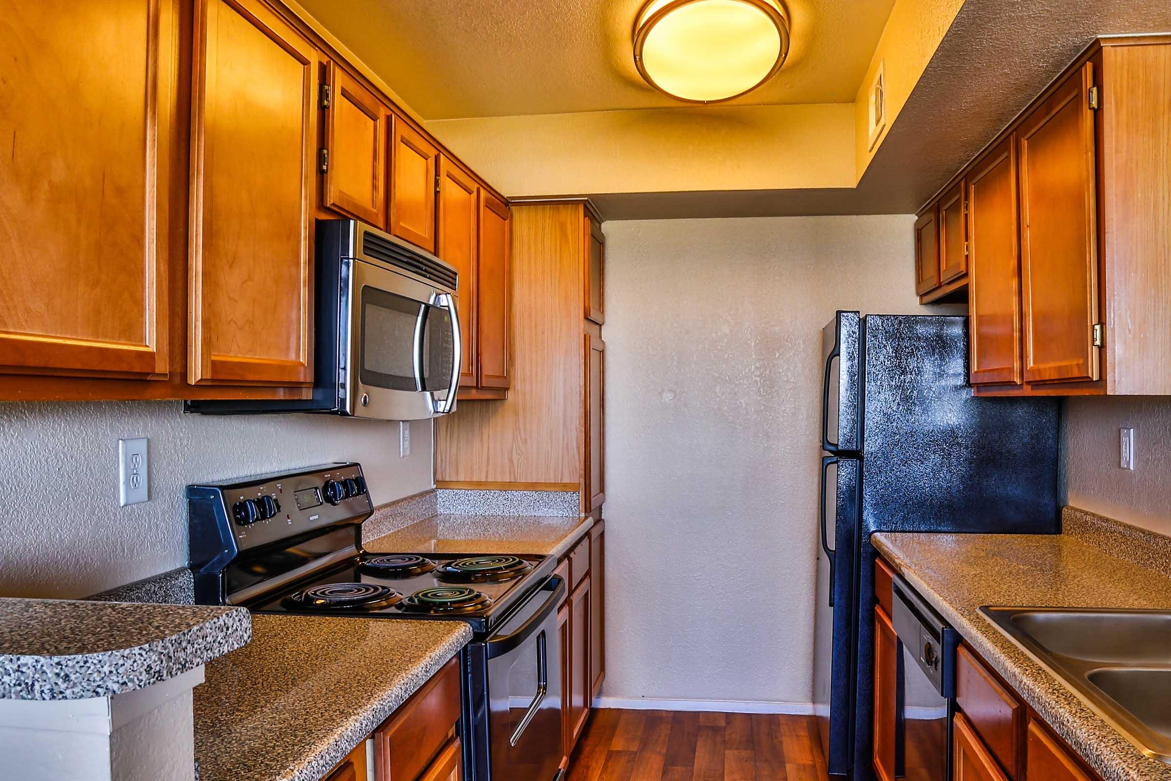 Modern kitchen featuring wooden cabinetry, stainless steel appliances, a black refrigerator, an electric stove with a built-in oven, and a stone countertop. The room has a warm ambiance with overhead lighting and a wood floor, creating a cozy and functional cooking space.