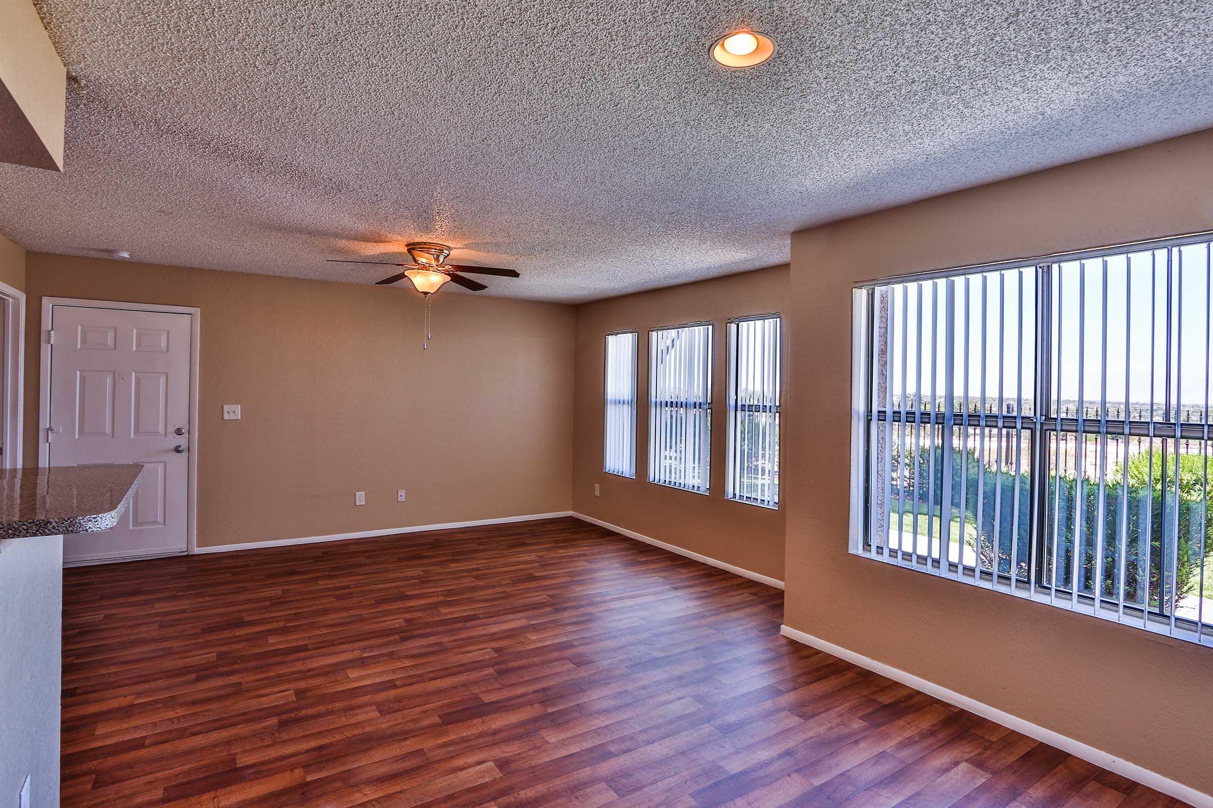 Spacious, well-lit living room with hardwood flooring, large windows featuring vertical blinds, a ceiling fan, and a neutral-colored walls. A view of greenery is visible through the windows, and a doorway leads to another room.