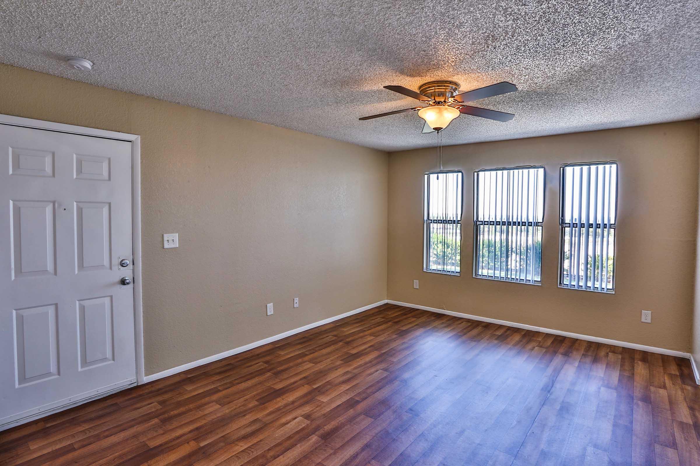 A bright, empty living room featuring a textured ceiling with a ceiling fan, large windows with vertical blinds, and wooden laminate flooring. The walls are painted a neutral color, and a front door with a small window is visible. The space is well-lit and suitable for furnishing.