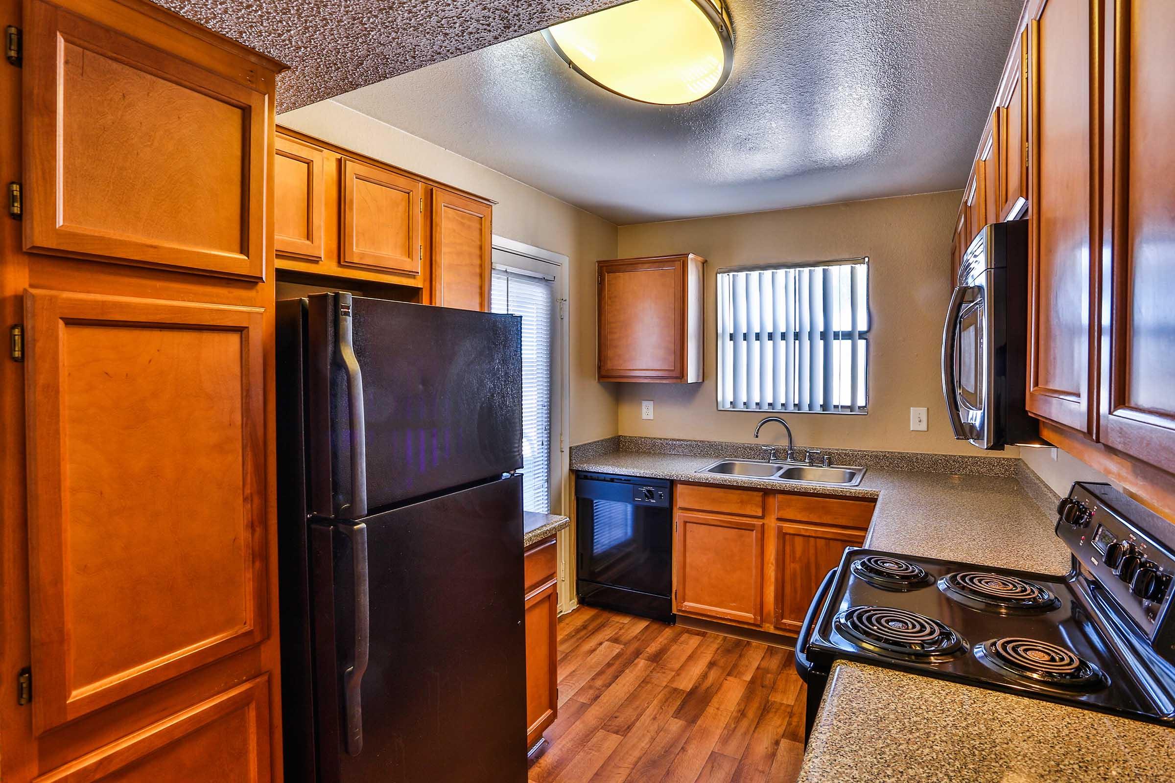 A modern kitchen with wooden cabinets, a black refrigerator, and stainless steel appliances. The countertop is a light gray, and there is a window with vertical blinds. The kitchen includes an oven, stovetop, microwave, and dishwasher, all arranged efficiently in a compact space with wooden flooring.