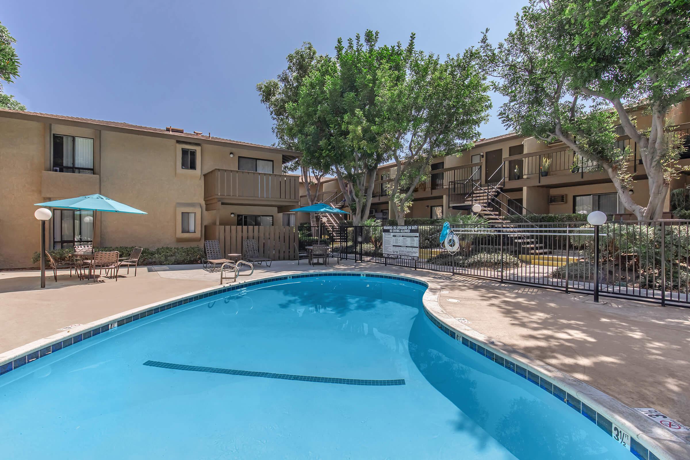 A clear blue pool surrounded by a sunlit courtyard with trees and lounge chairs. Apartment buildings are in the background, and umbrellas provide shade. A few steps lead to the pool area, creating a relaxing atmosphere. The scene is bright and inviting, perfect for outdoor leisure.