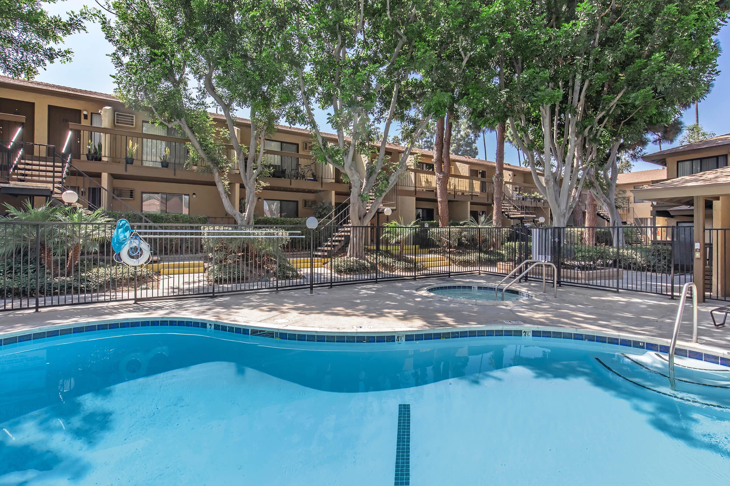 A clear swimming pool surrounded by a fenced area with lounge chairs and palm trees. There are buildings in the background, featuring staircases leading to upper levels, set in a sunny outdoor environment.