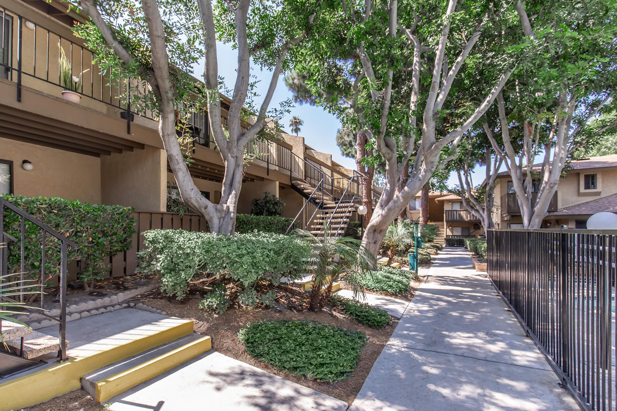 A pathway lined with greenery leads to a set of two-story apartment buildings. The area features trees and well-maintained shrubs, with stairs visible leading up to the entrance of the buildings. The setting appears peaceful and residential, suitable for living.