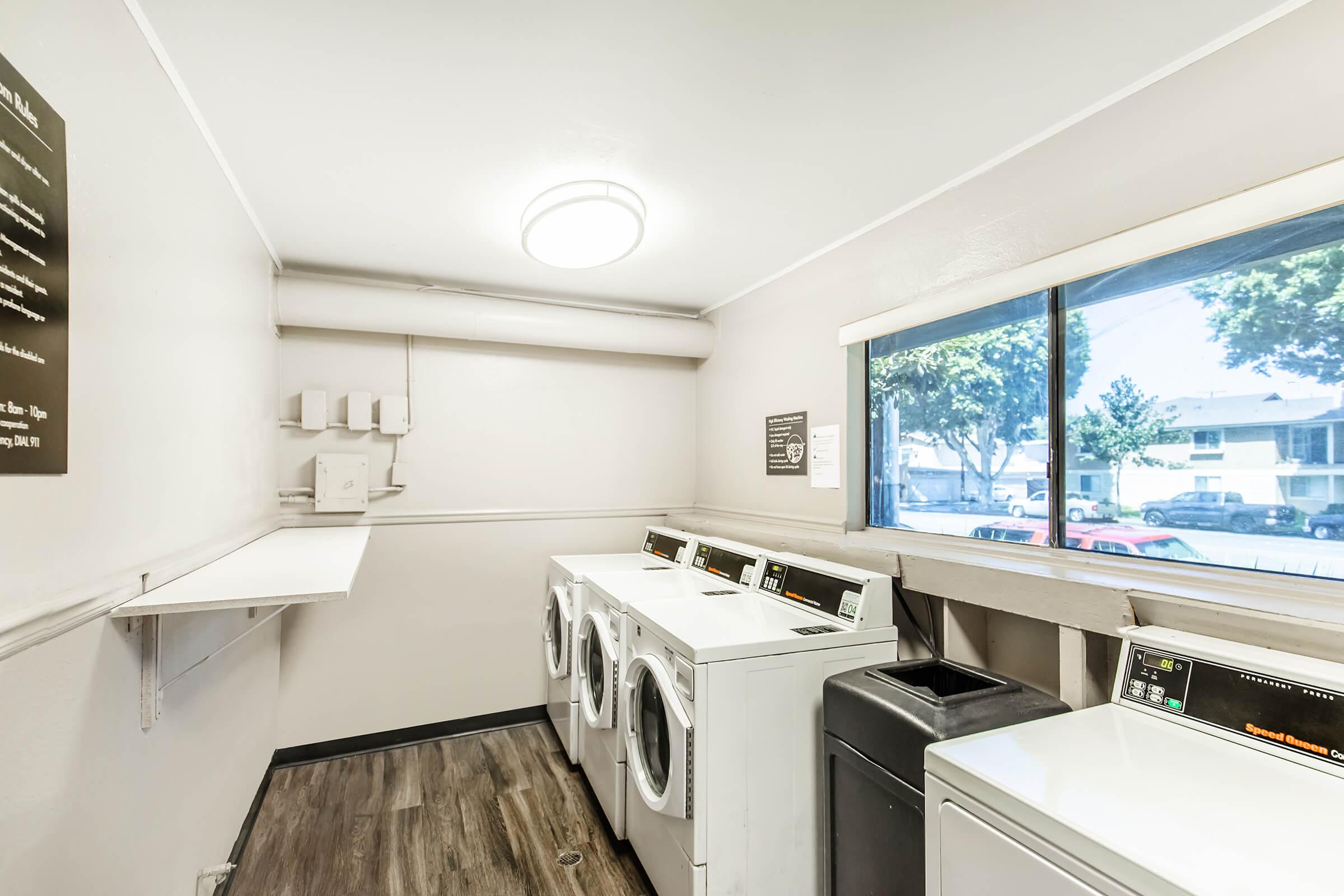 A clean, well-lit laundry room featuring several white washing machines and a dryer. There's a window providing natural light and a shelf attached to the wall. The space has light-colored walls and laminate flooring, with a modern ceiling light fixture.