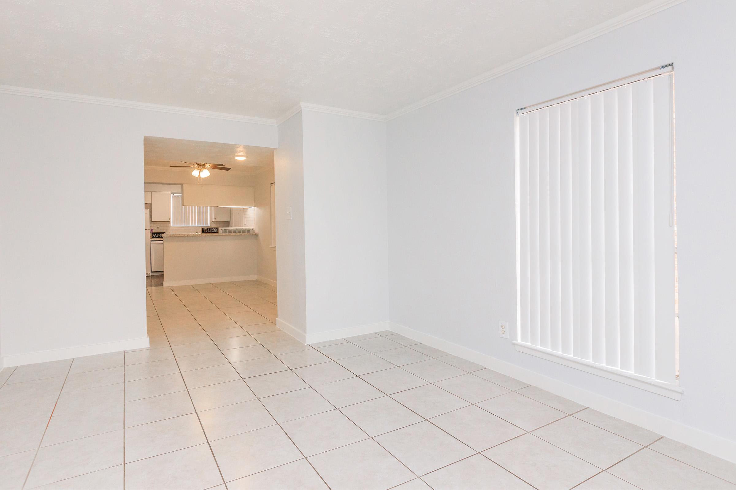 A spacious, well-lit room with light-colored walls and floor tiles. There is an open doorway leading to another area of the home, which appears to have a kitchen. A window with vertical blinds is visible on one wall, adding natural light to the space. The overall ambiance is clean and modern.