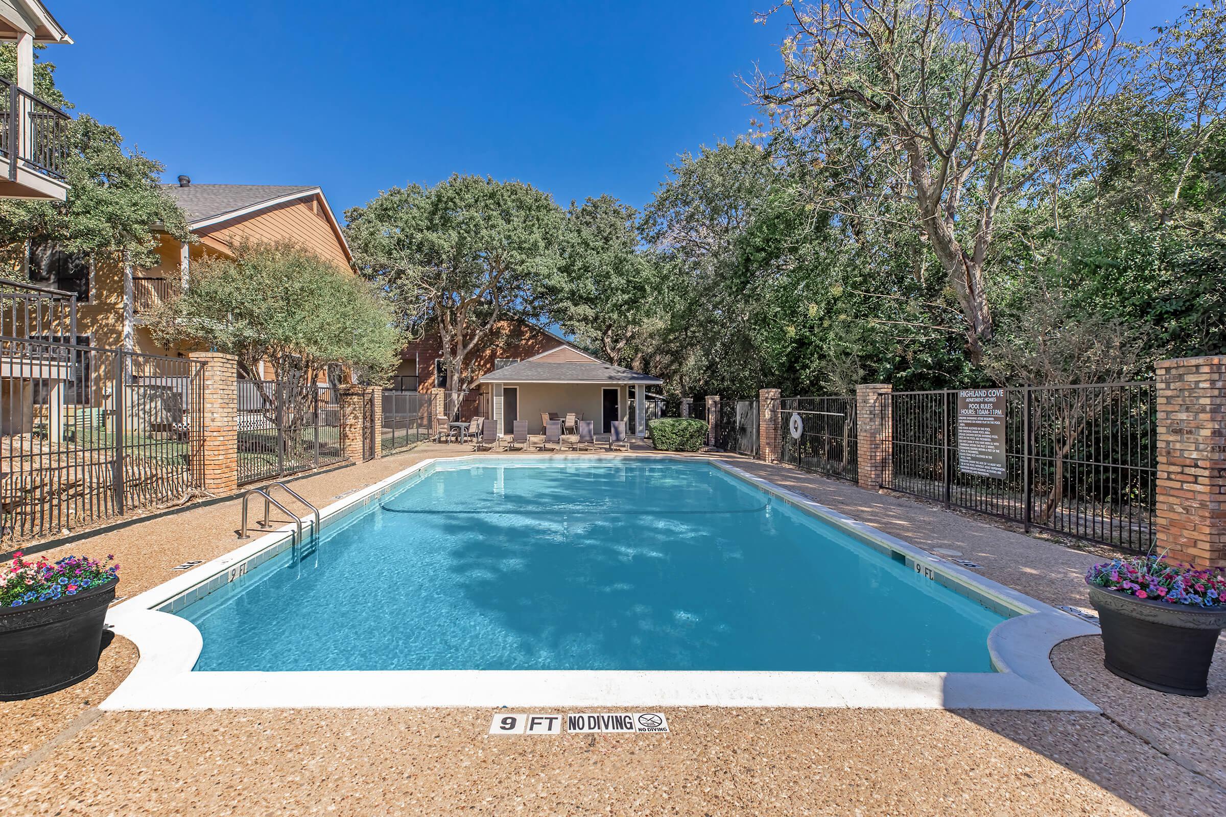 A clear swimming pool surrounded by a fenced area, with a shallow end marked as "9 FT NO DIVING." Lush greenery and trees provide a scenic backdrop, while a covered seating area is visible in the background. Colorful flower pots are placed at the pool's edge, adding a decorative touch.