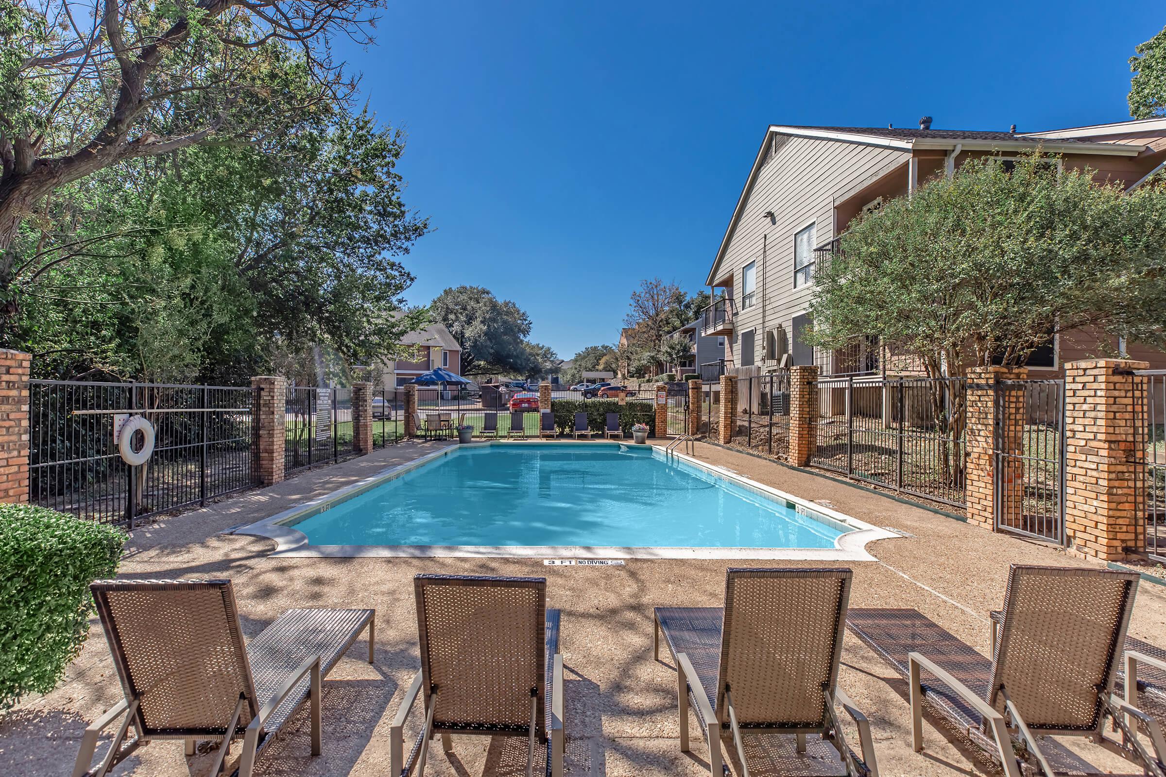 A view of a residential pool area featuring a clear blue swimming pool surrounded by lounge chairs. The space is enclosed by a fence and has lush greenery and trees nearby. Several apartment buildings are visible in the background under a clear blue sky.