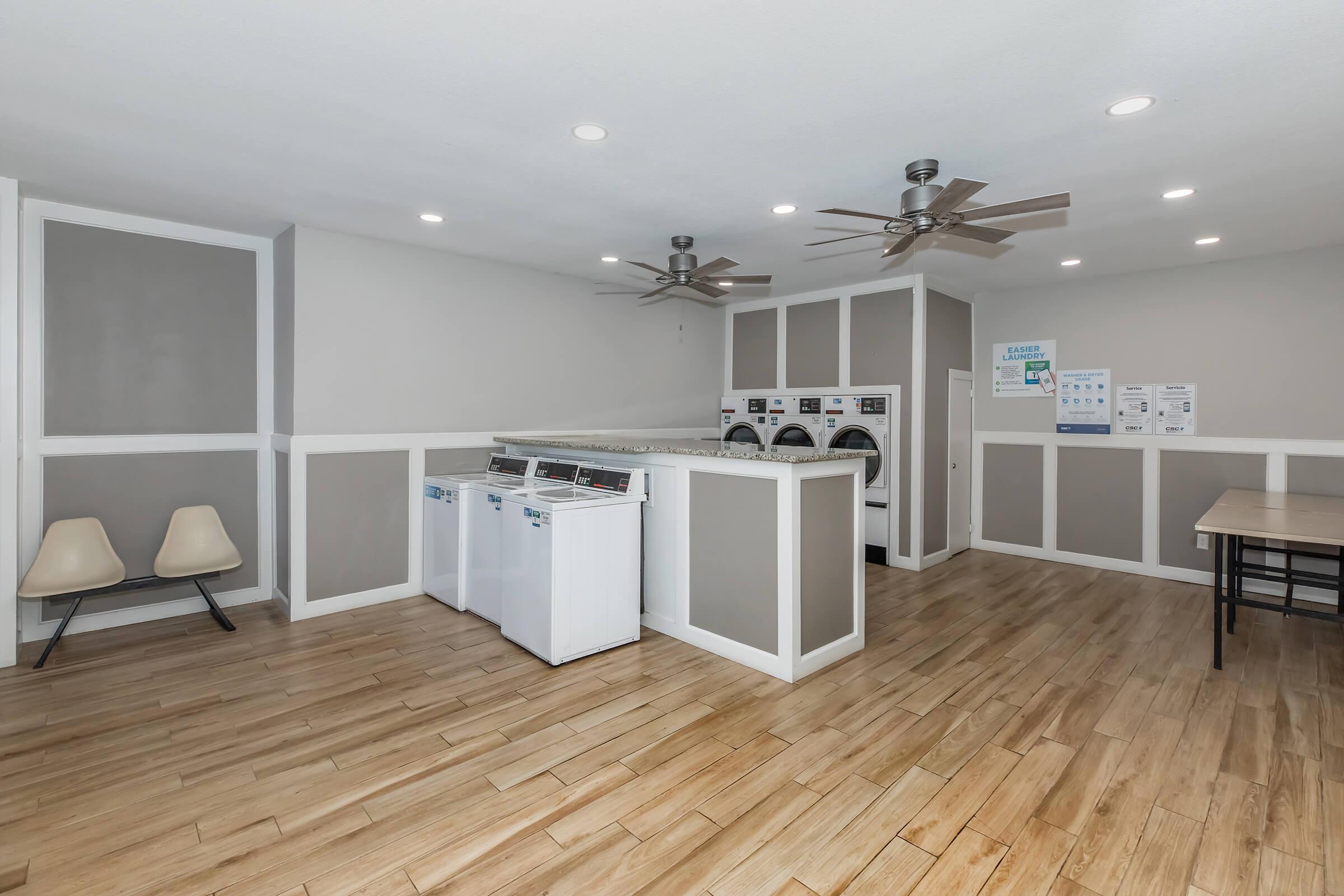 A tidy laundry room featuring multiple white washing machines and dryers against a light grey wall. The space includes modern lighting, ceiling fans, and wooden flooring. There are two beige chairs on one side and a large table on the opposite side, creating a functional and comfortable environment for doing laundry.