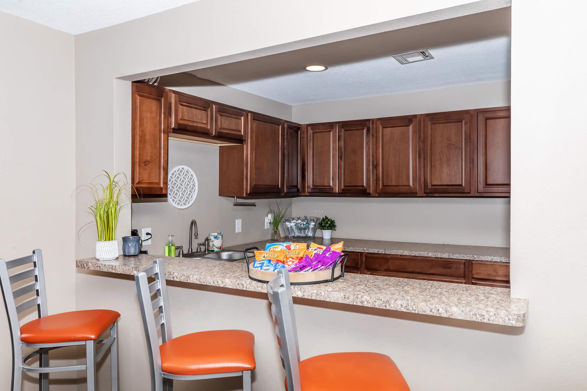 A modern kitchen featuring dark wooden cabinetry, a granite countertop with bar seating, and orange upholstered stools. There are various kitchen appliances, decorative plants, and a basket of snacks on the counter. The walls are painted a neutral color, creating a warm and inviting atmosphere.