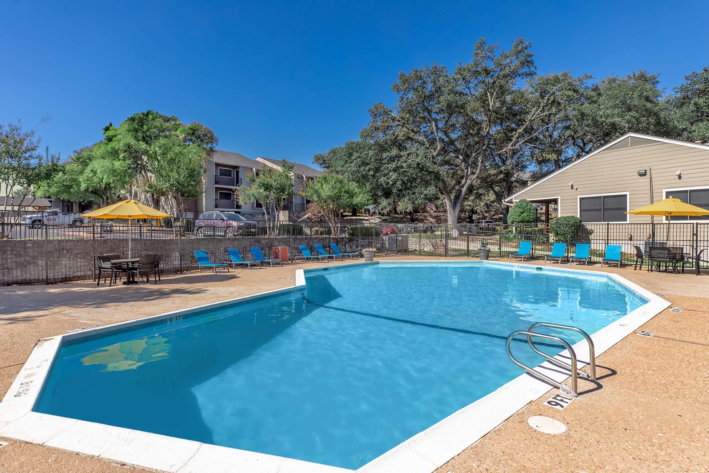 A bright, inviting swimming pool surrounded by lounge chairs in a sunny outdoor area. There are umbrellas for shade, and a nearby building with trees and landscaping in the background. The scene conveys a relaxing atmosphere perfect for leisure and recreation.