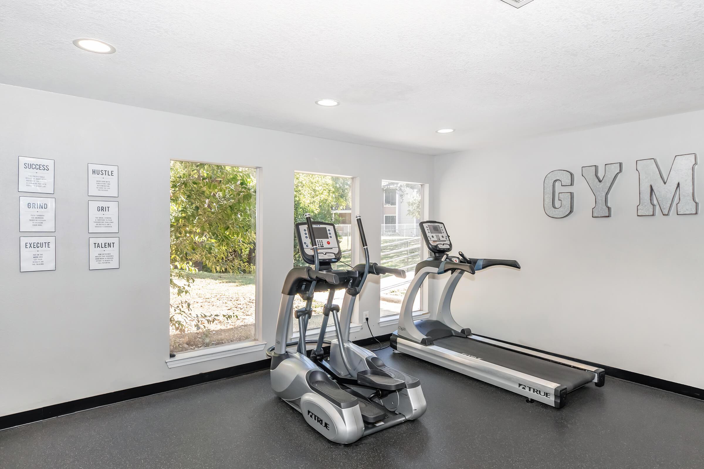 A modern gym interior featuring two pieces of exercise equipment: an elliptical machine and a treadmill. The walls are adorned with motivational quotes framed on the left, and large windows allow natural light to brighten the space with a view of greenery outside. The word "GYM" is displayed prominently on one wall.