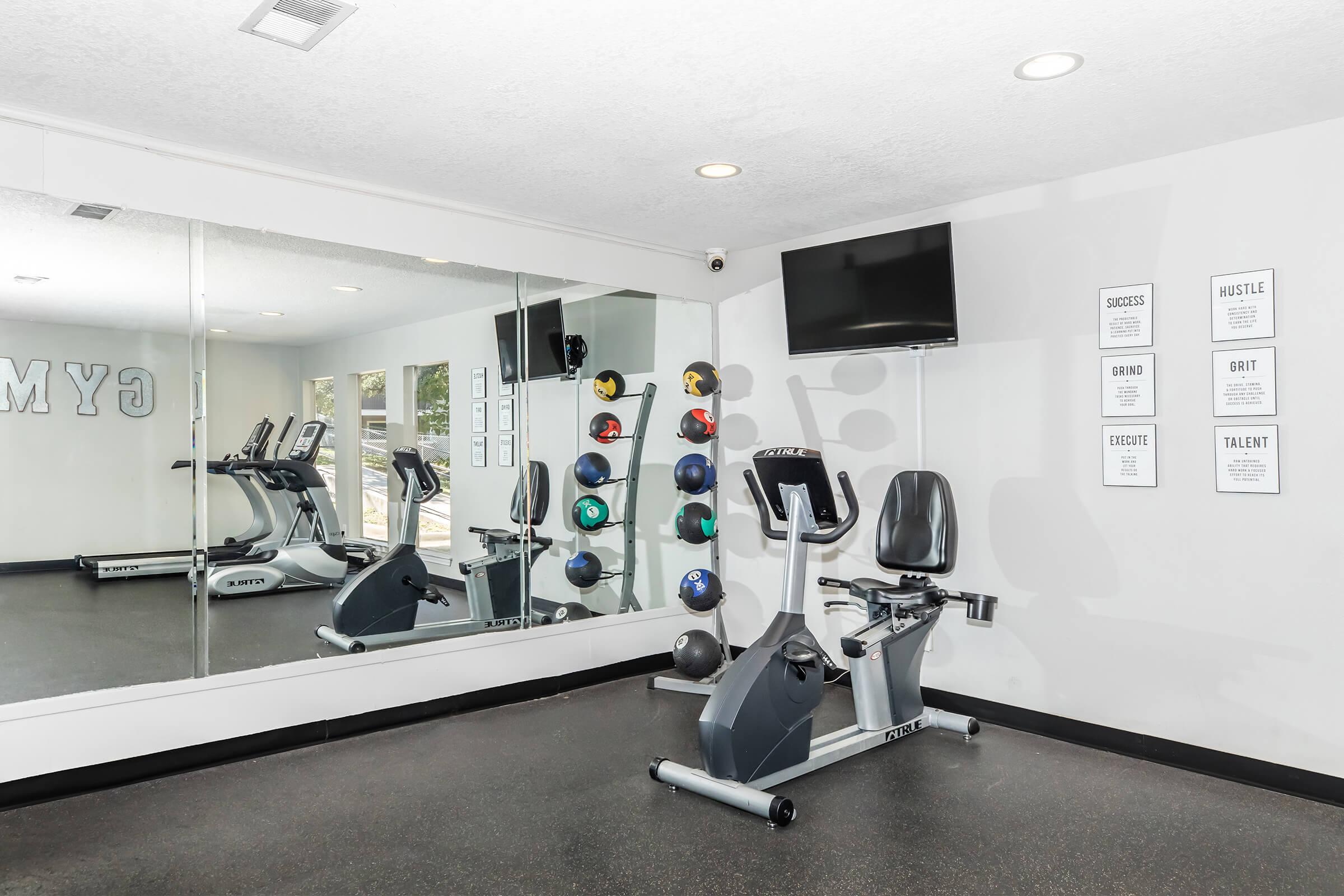 A modern gym interior featuring exercise equipment, including a stationary bike and treadmills. The wall mirrors reflect the space, while colorful dumbbells are neatly arranged on a rack. Inspirational posters hang on the wall, promoting motivation and fitness. Soft lighting enhances the inviting atmosphere.