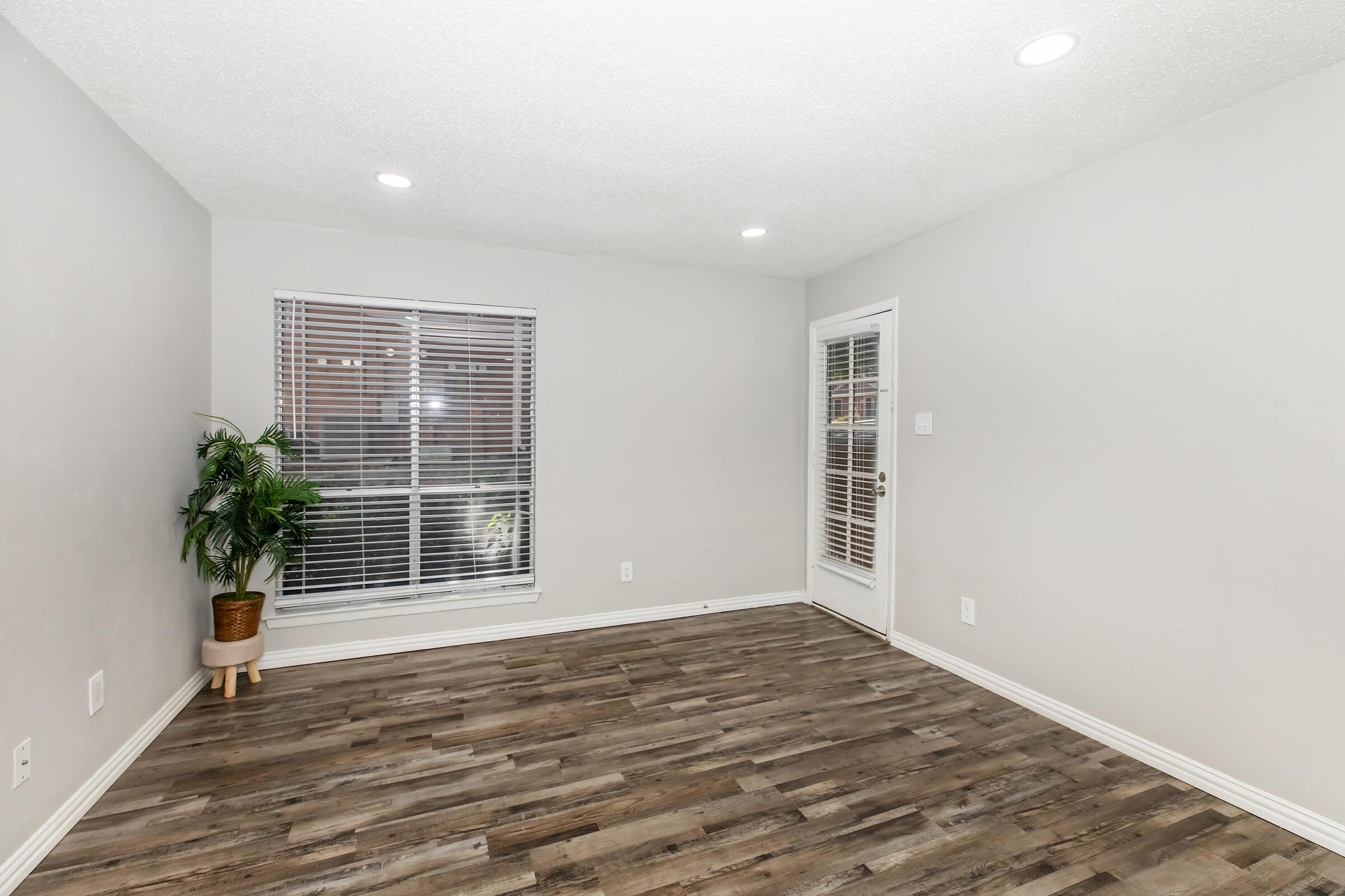 A spacious, unfurnished room featuring light gray walls, laminate flooring, and a large window with blinds. A potted plant sits in the corner, and there is a door leading outside. The room is well-lit with recessed lighting on the ceiling.