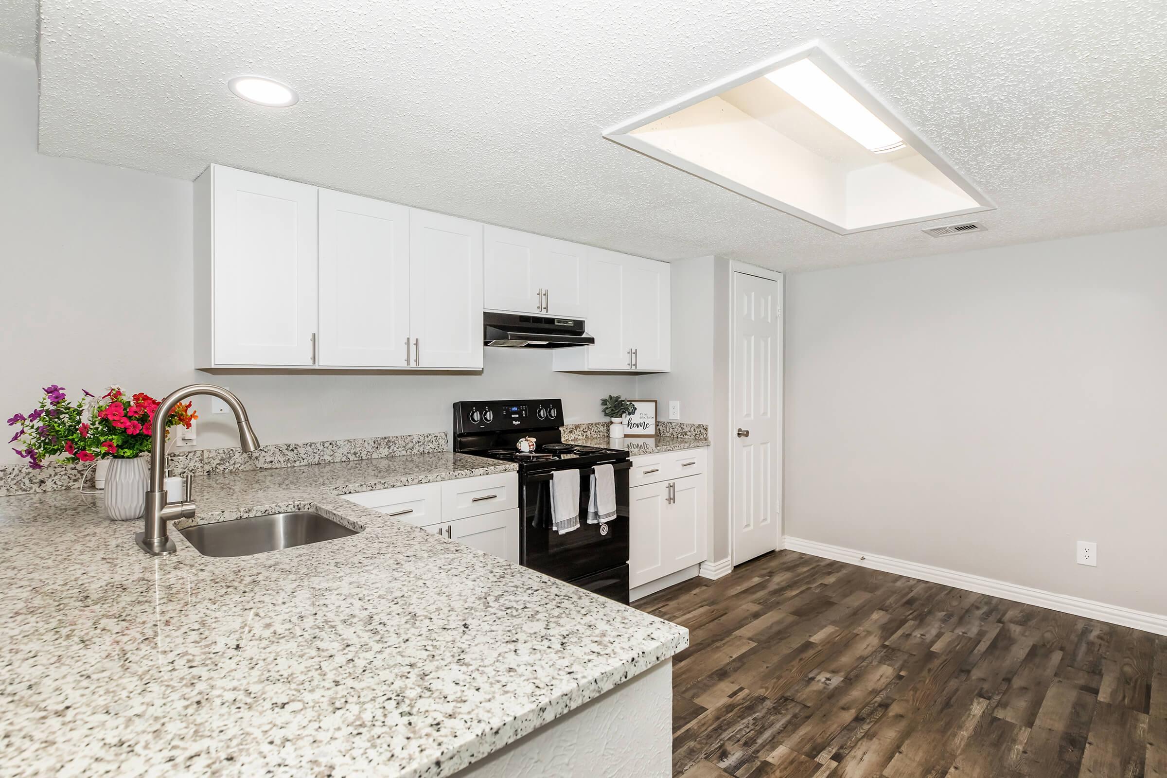 Modern kitchen featuring white cabinets, a granite countertop, and stainless steel appliances. There's an overhead light fixture and a sink with a sleek faucet. Fresh flowers are arranged on the counter, and the flooring is a warm wood finish.