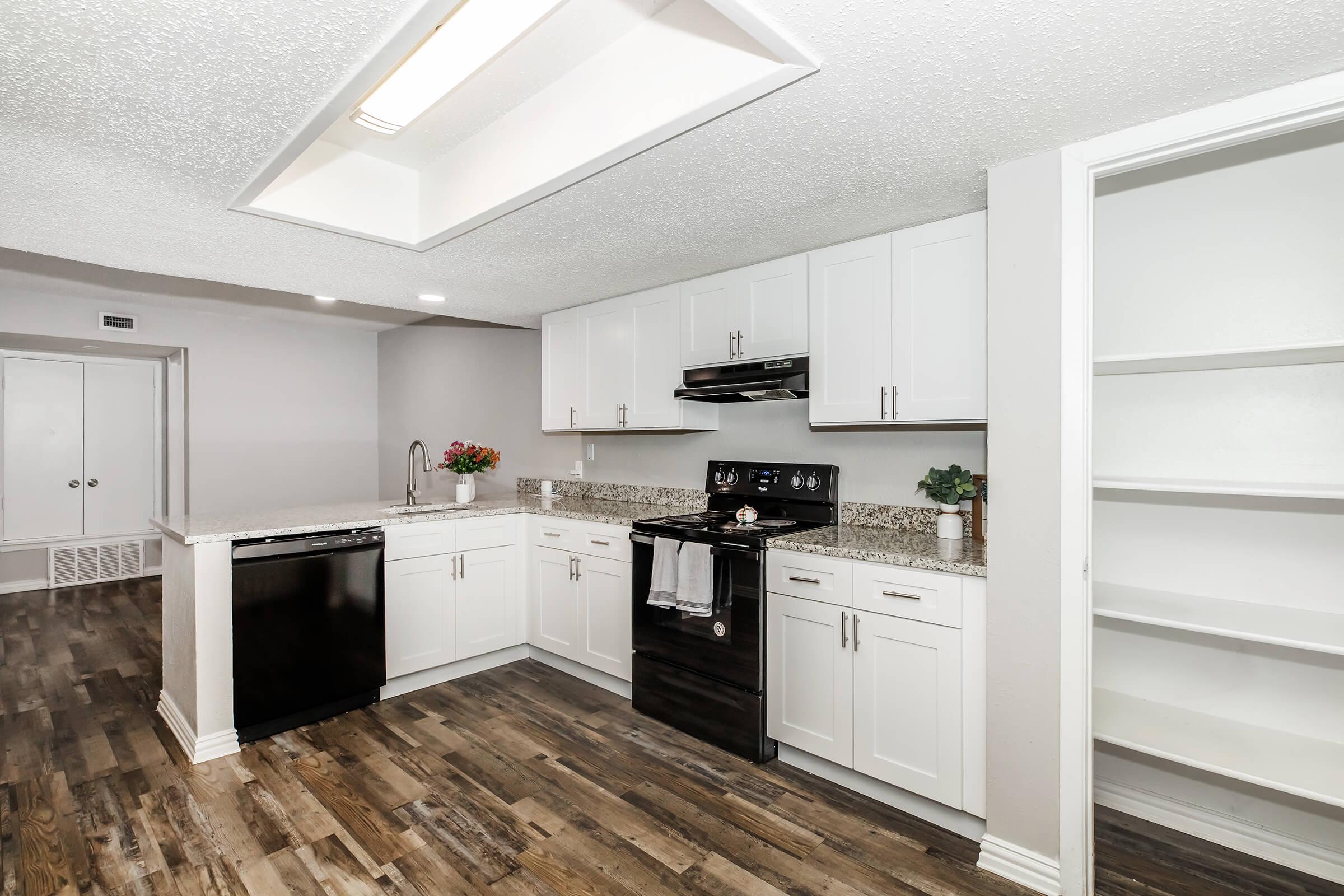 Modern kitchen featuring white cabinets, granite countertops, and stainless steel appliances including a dishwasher and stove. The space has wood-like flooring and a large storage pantry. Bright lighting enhances the open layout, with plants adding a decorative touch.