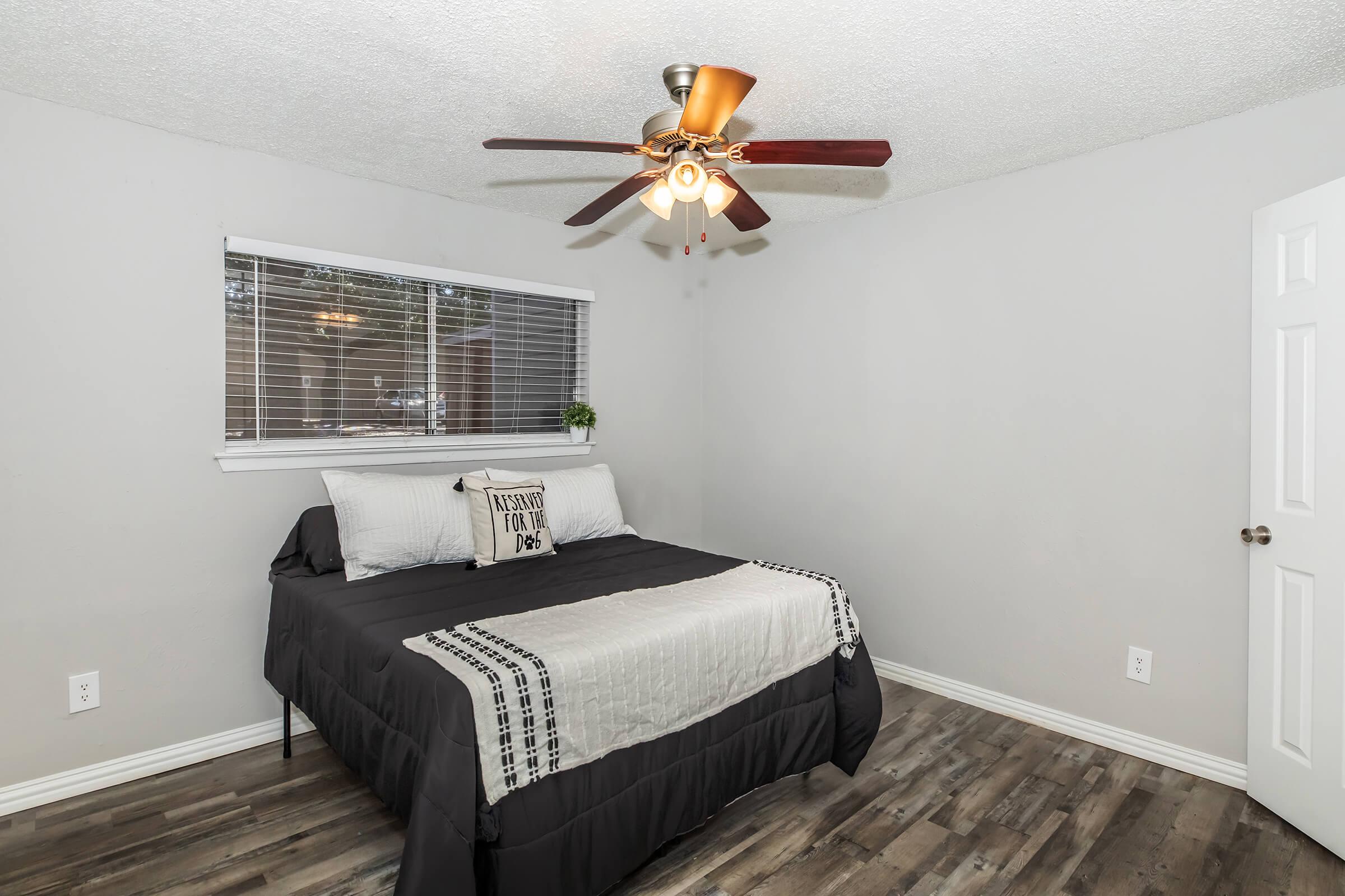 A cozy bedroom featuring a queen-sized bed with gray bedding and a decorative pillow. There's a ceiling fan with light fixtures, a window with blinds, and light-colored walls. The flooring is dark wood, and a door is visible on the right side of the image.