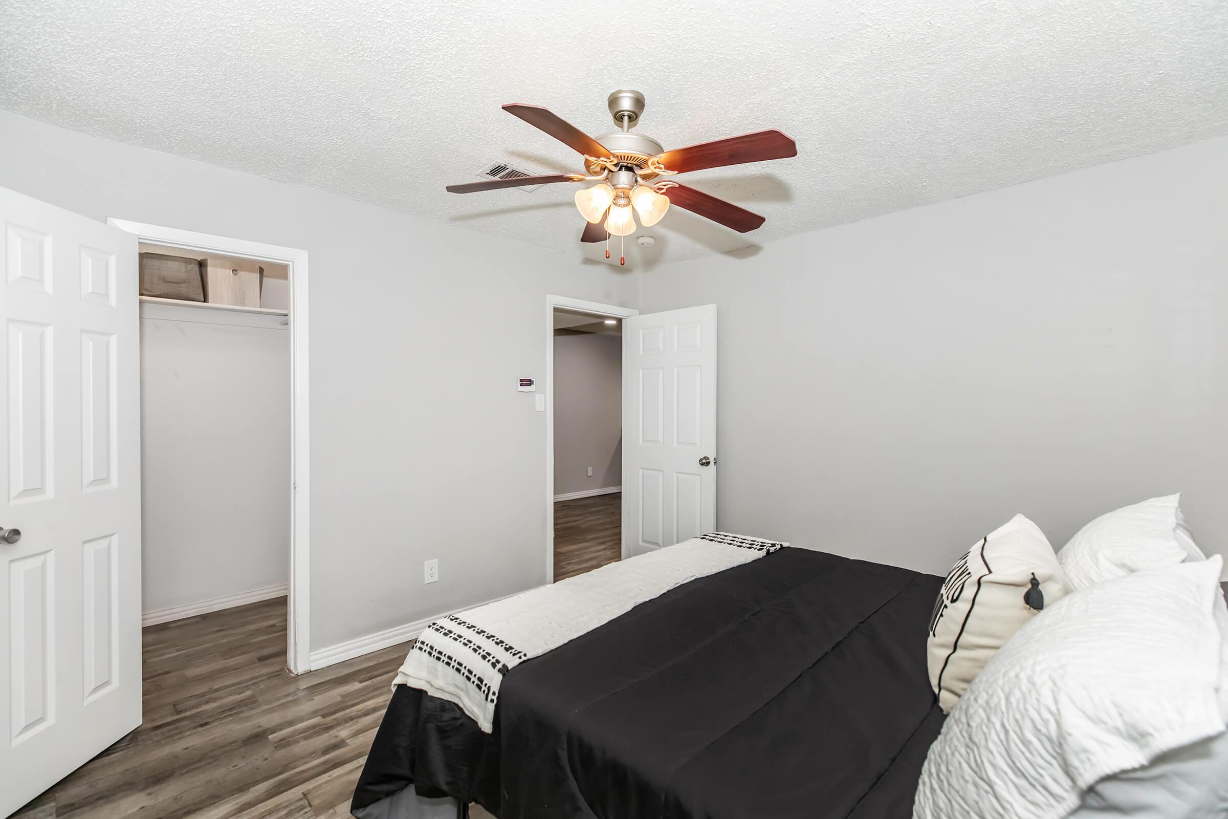 Interior of a bedroom featuring a ceiling fan with light, a neatly made bed with black and white bedding, and wooden flooring. Two doors are visible, leading to a closet and another room. The walls are painted in a light color, contributing to a bright, spacious feel.