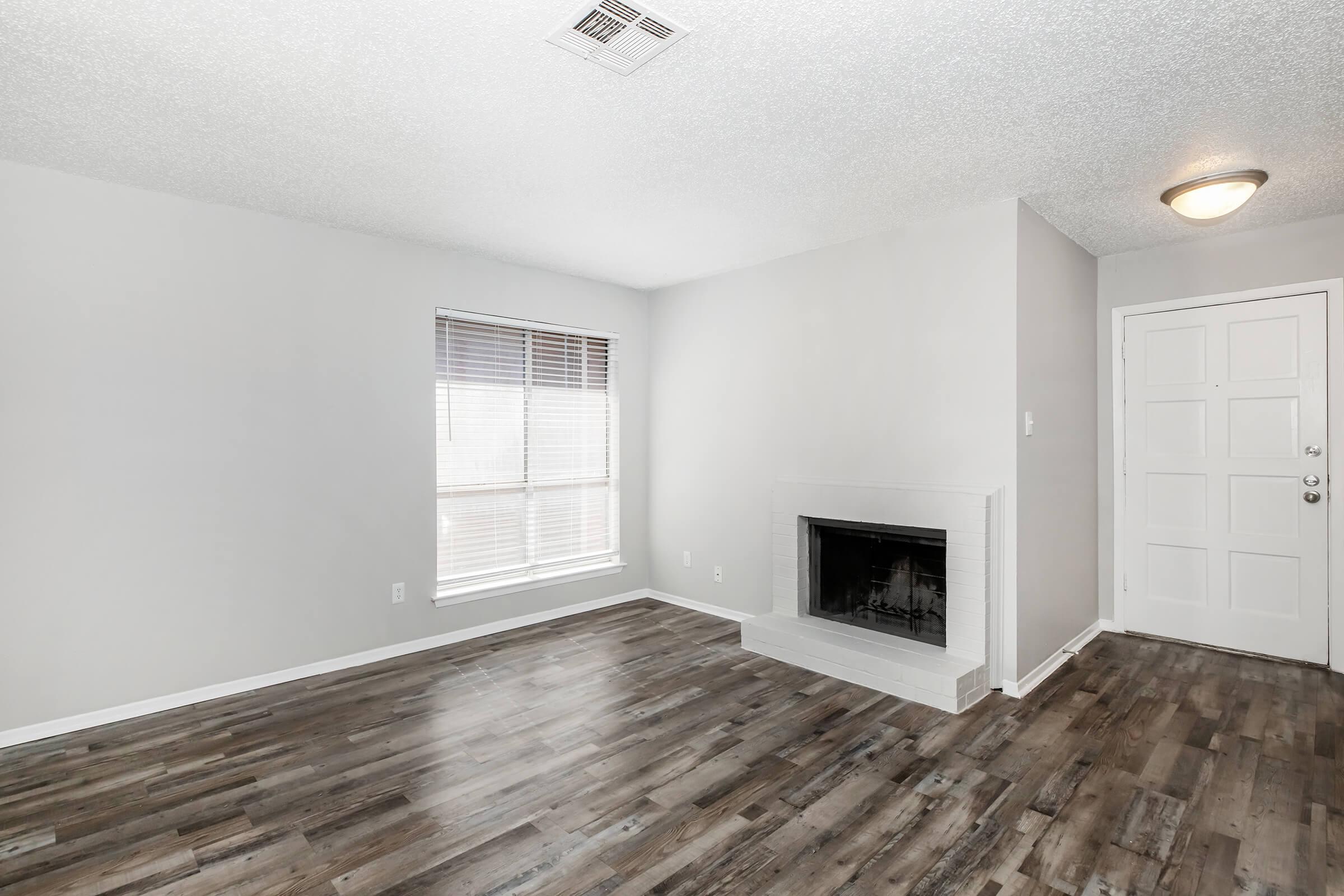 A spacious, empty living room featuring a fireplace, large window with blinds, and gray walls. The floor is made of dark wood laminate. A front door is visible on the right side of the image, adding to the open and airy feel of the room. Lights are on the ceiling, giving a warm ambiance.