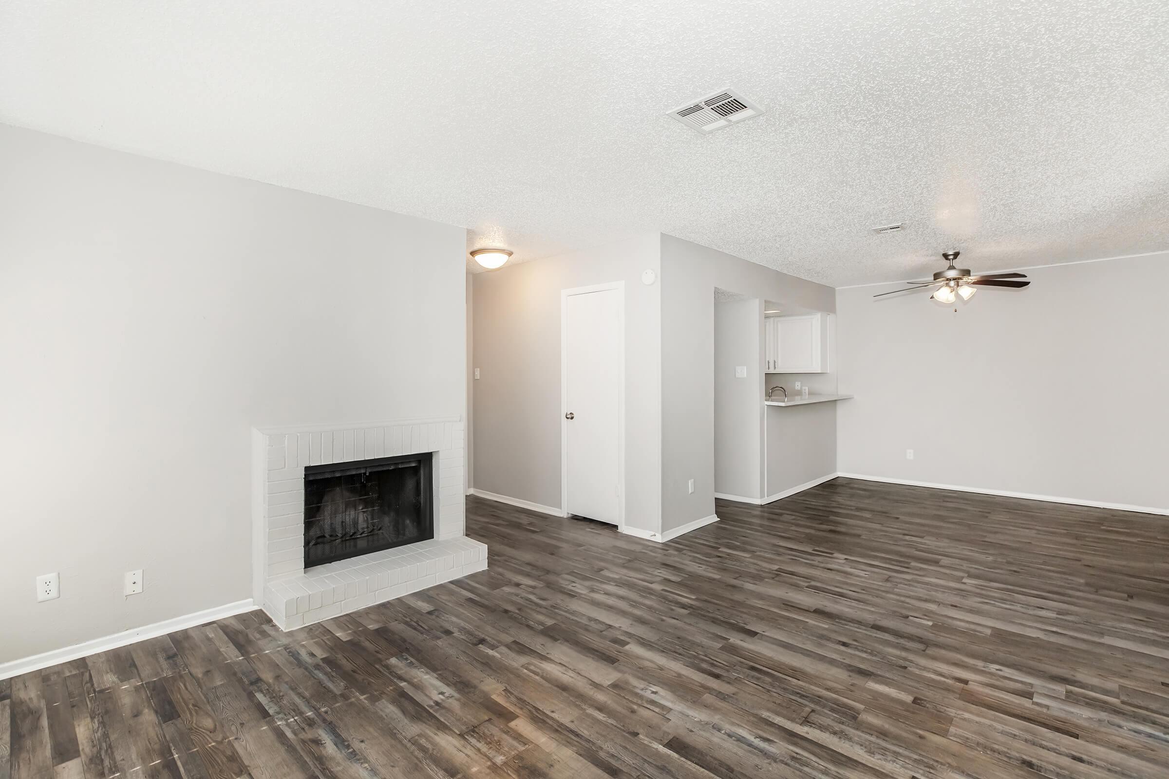 A spacious living area featuring a white brick fireplace, light gray walls, and wooden floors. A ceiling fan is visible, and there is an entryway leading to a kitchen area with a bar. The overall ambiance is bright and inviting, ideal for relaxation and entertaining.