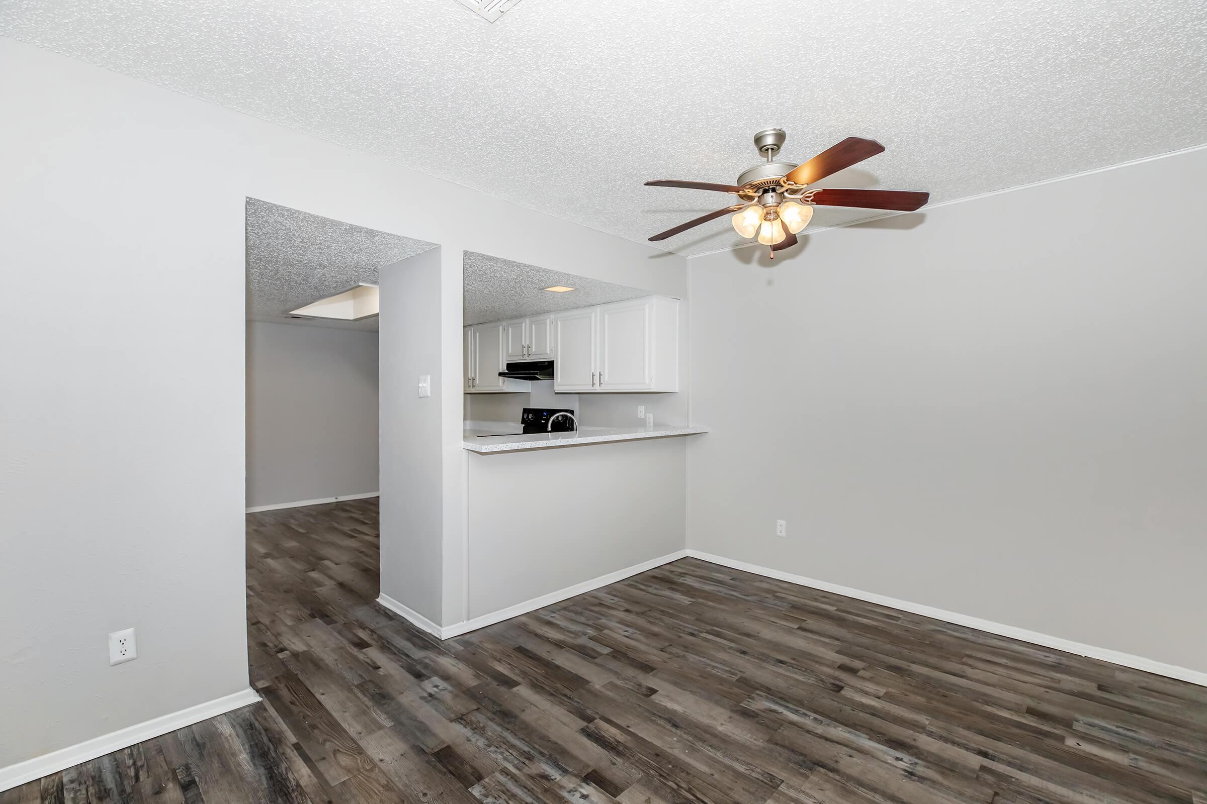 A spacious, well-lit living area featuring a ceiling fan, light wood flooring, and neutral-colored walls. A partial wall opens to a kitchen with white cabinets and a countertop. The room has a warm, inviting atmosphere, ideal for relaxation or socializing.